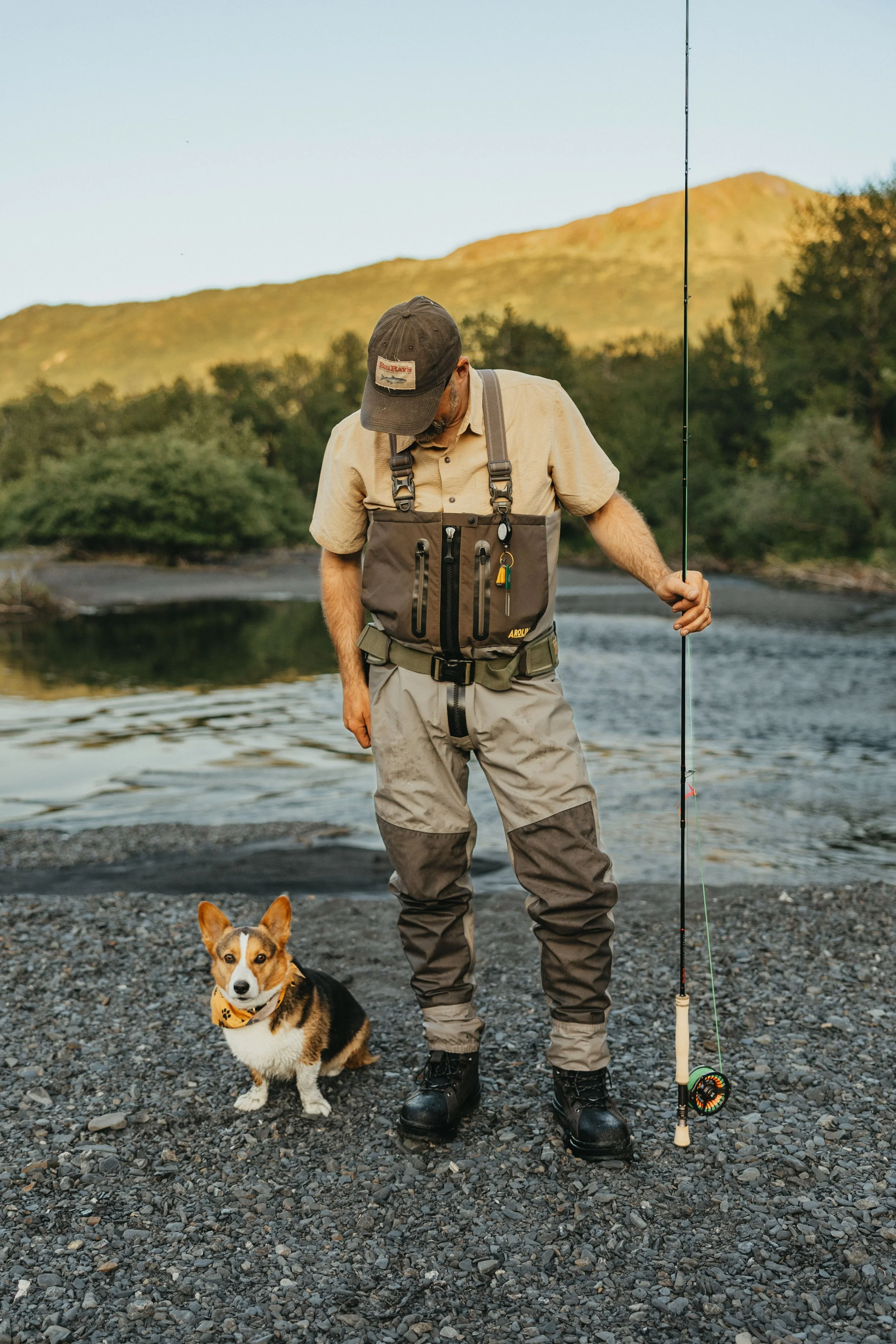 Man in Arolik fishing waders standing on a rocky Alaska riverbank with a dog, holding a fly rod during golden hour outdoor fishing.