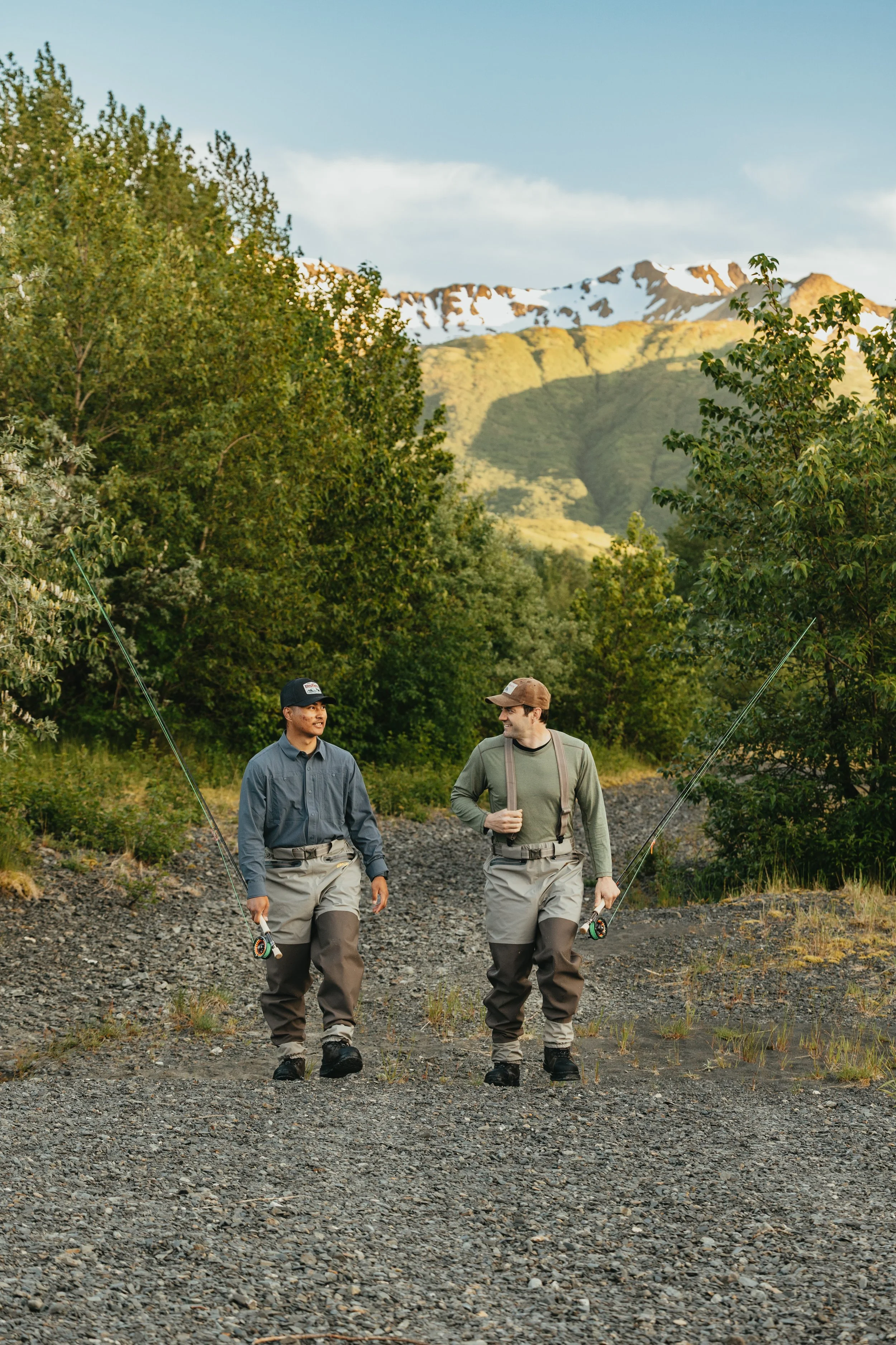 Two anglers walking along an Alaska trail in Arolik waders with fishing rods, surrounded by mountains and forest.