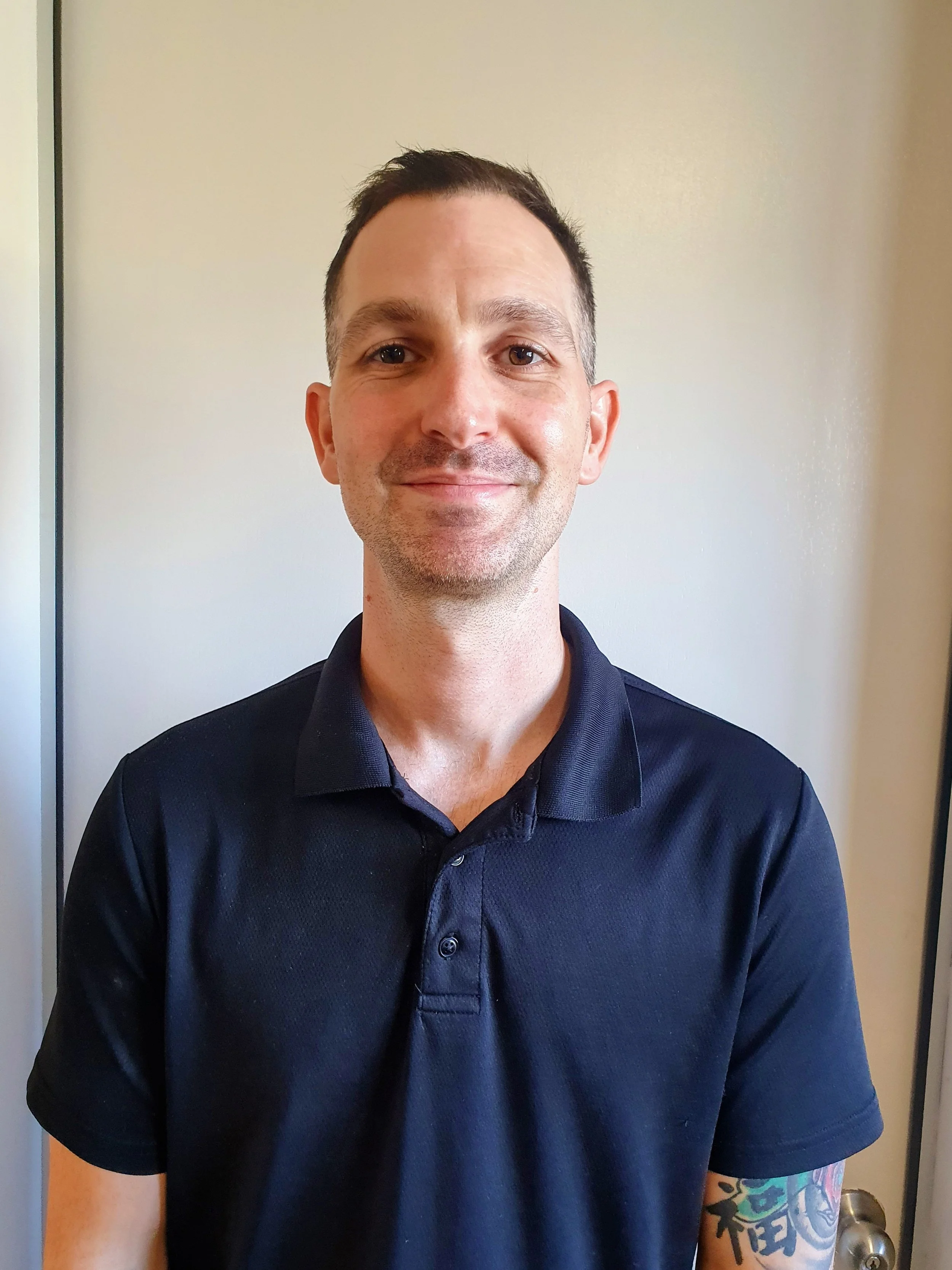 A man with short dark hair, wearing a black polo shirt, smiling, standing in front of a plain wall. Massage. Remedial Massage. Sports Massage. Bribie Island