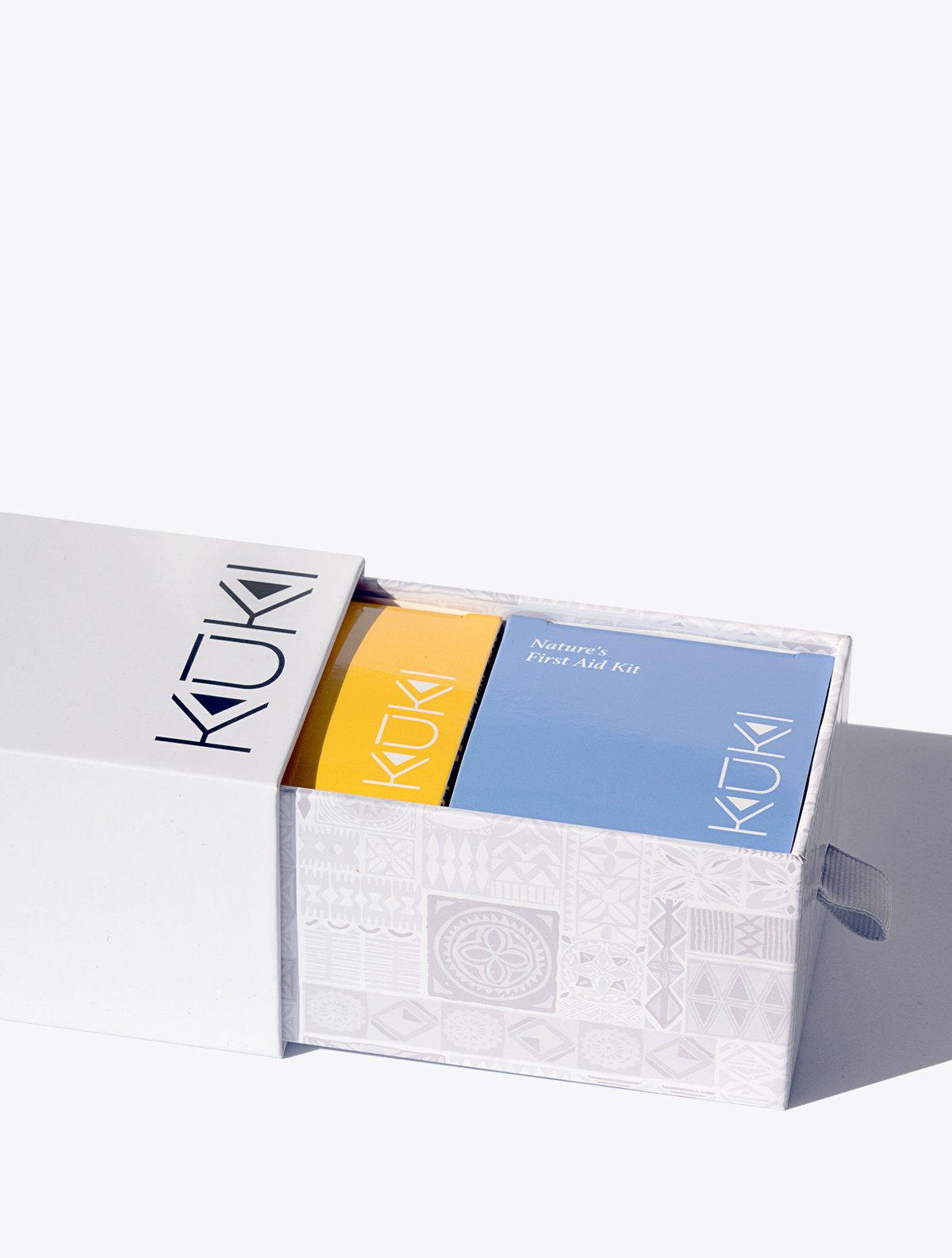 Open gift box with KUKI branded packaging, showing two colorful product boxes inside, one yellow and one blue, labeled 'Nature’s First Aid Kit'.