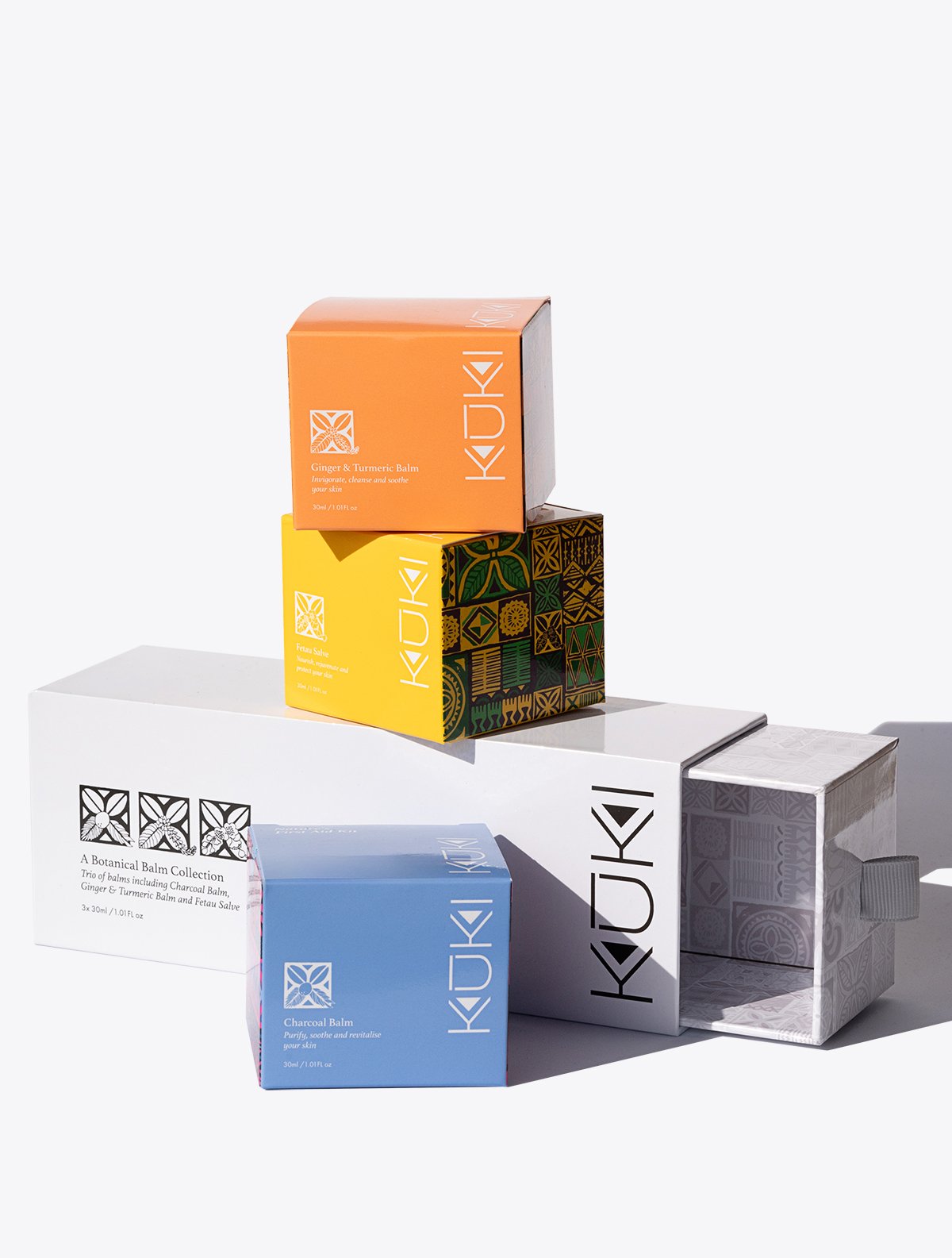 KUKI botanical balm packaging, featuring colorful boxes for Charcoal Balm, Ginger & Turmeric Balm, and other products, stacked together.