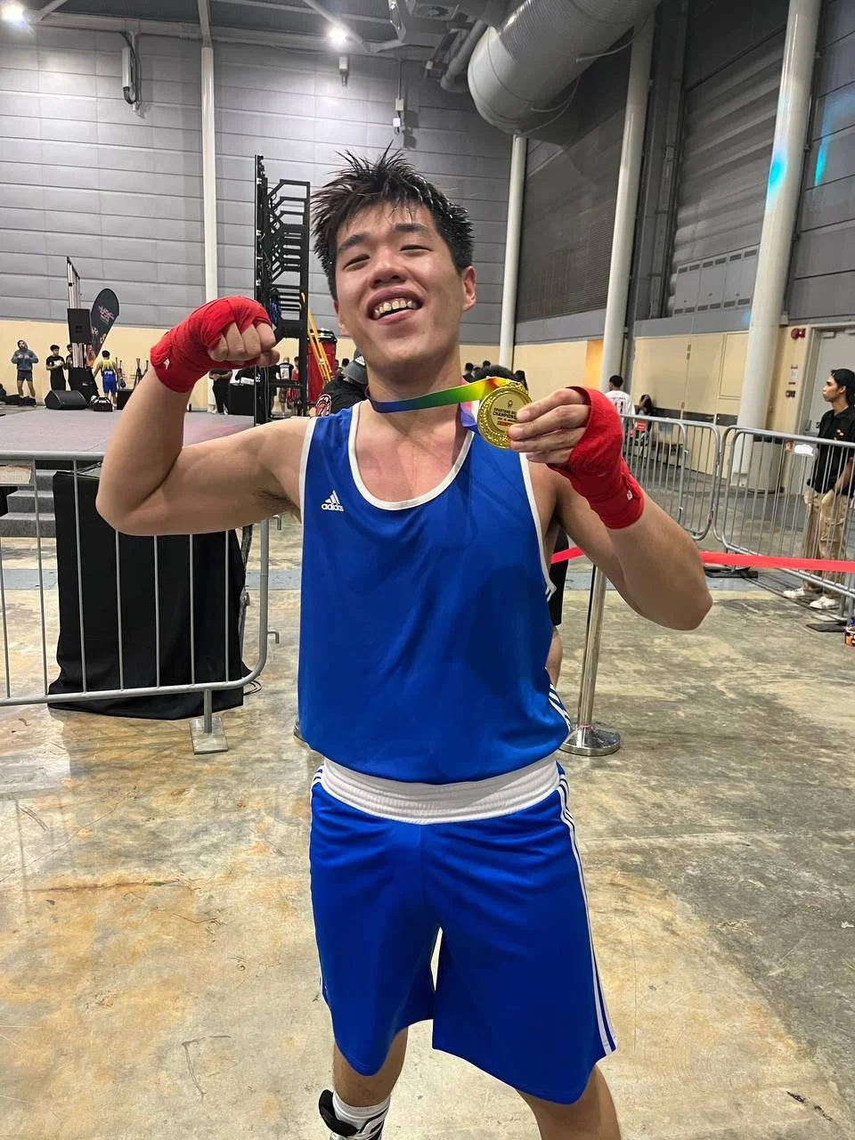 A young male boxer in blue sportswear, wearing red hand wraps, proudly displaying a gold medal at a sports event. He is smiling and flexing his right arm, with event equipment and other people in the background.