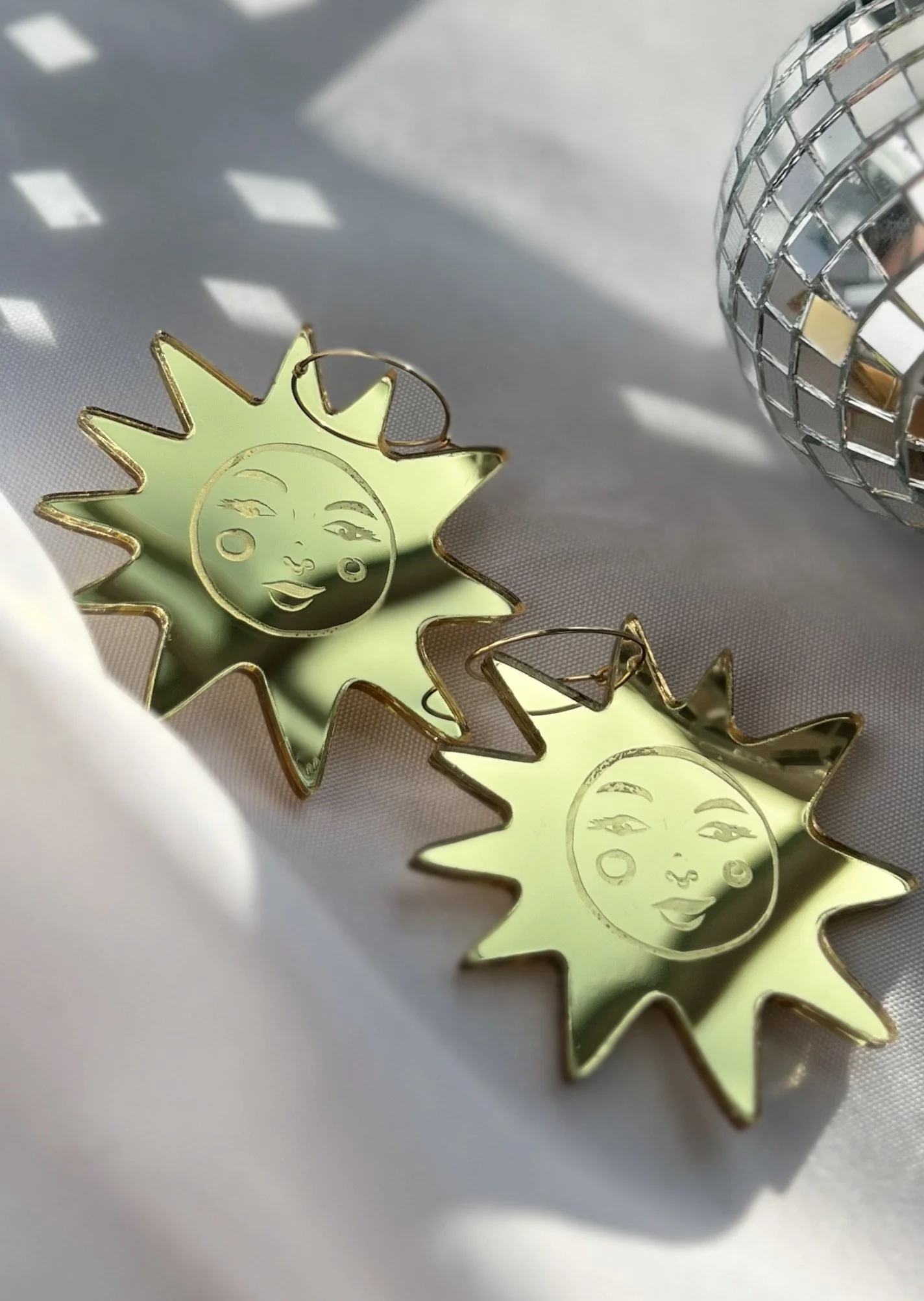 Pair of metallic earrings shaped like the sun with a face, lying on white fabric next to a small disco ball reflection.