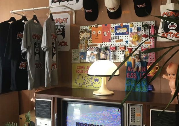 Display of hanging t-shirts and caps, a wooden table with a retro TV, a lamp, and various colorful posters and toys in the background.