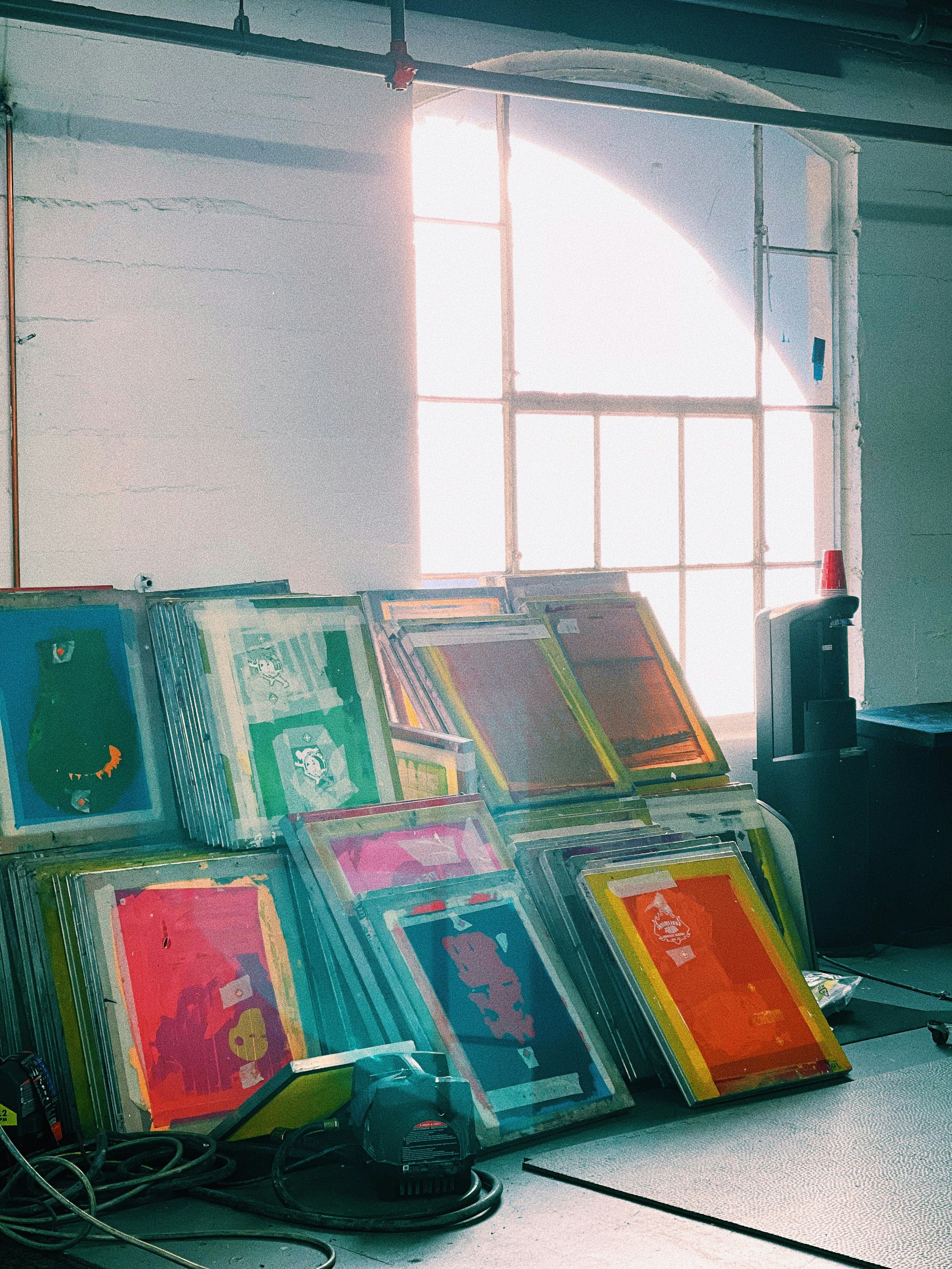 A stack of colorful framed artworks leaning against a wall near a large window with sunlight coming through, with some equipment and cables on the floor.