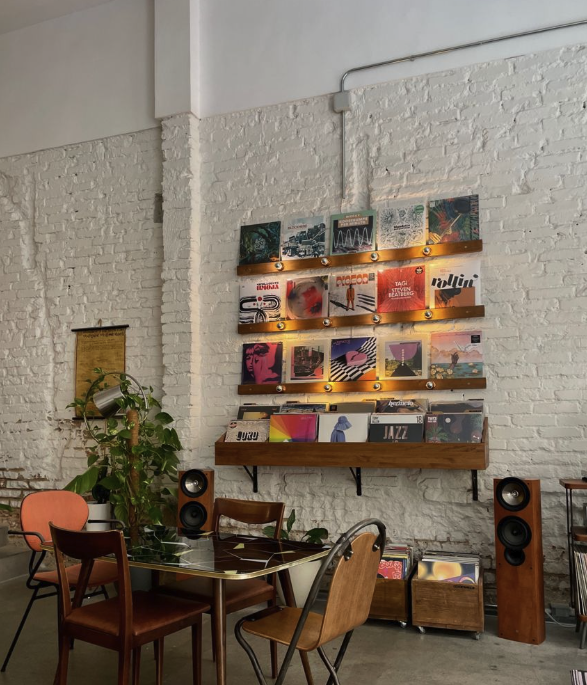 Indoor space with white brick wall decorated with vinyl records and artwork, a wooden shelf, potted plant, and audio speakers, with a table and chairs for seating.
