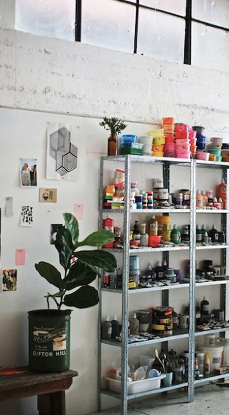 Shelves filled with various art supplies, including paint jars, brushes, and containers, in an artist's studio with a white brick wall and a large window.