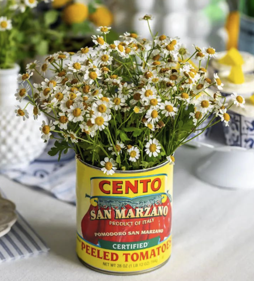 A bouquet of daisies in a yellow can labeled 'Cento San Marzano Peeled Tomatoes' on a table with a white tablecloth, with other decorative items in the background.