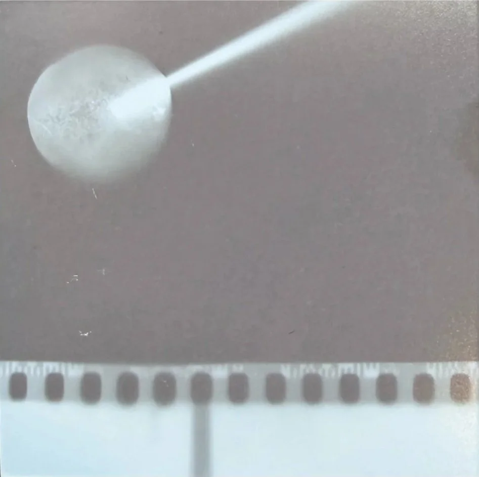 A photogram. The background is dark beige-purple. A circle, looking moon-like, sits at the top left being punctured by a thin white stick. The bottom is lined with a strip of 35mm film.