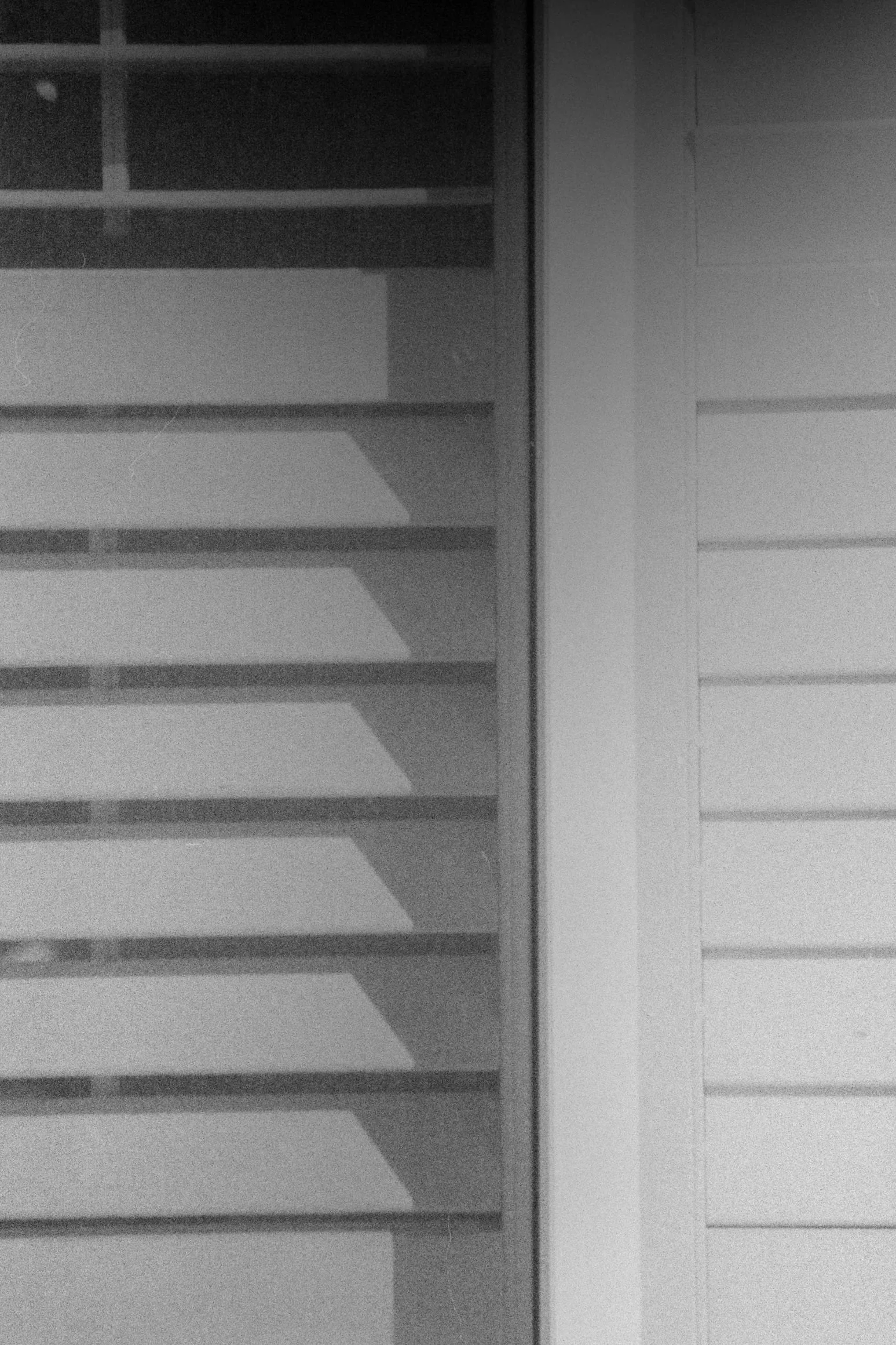 A hazy eco processed black & white 35mm film image shows window blinds from the outside. they are mostly shut, apart from a bit at the top right.