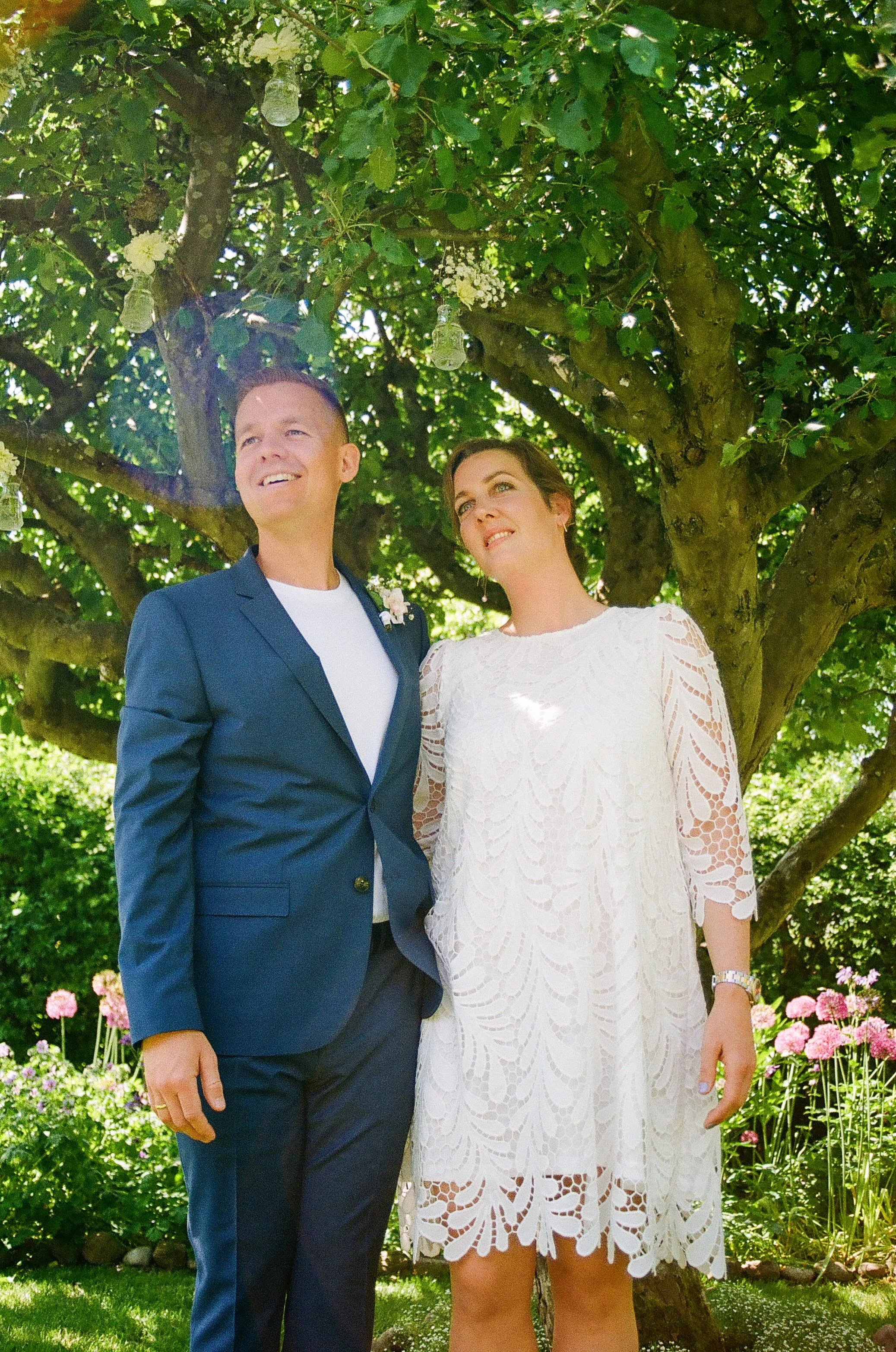 Couple posing outdoors under a tree, man in blue suit, woman in white lace dress.