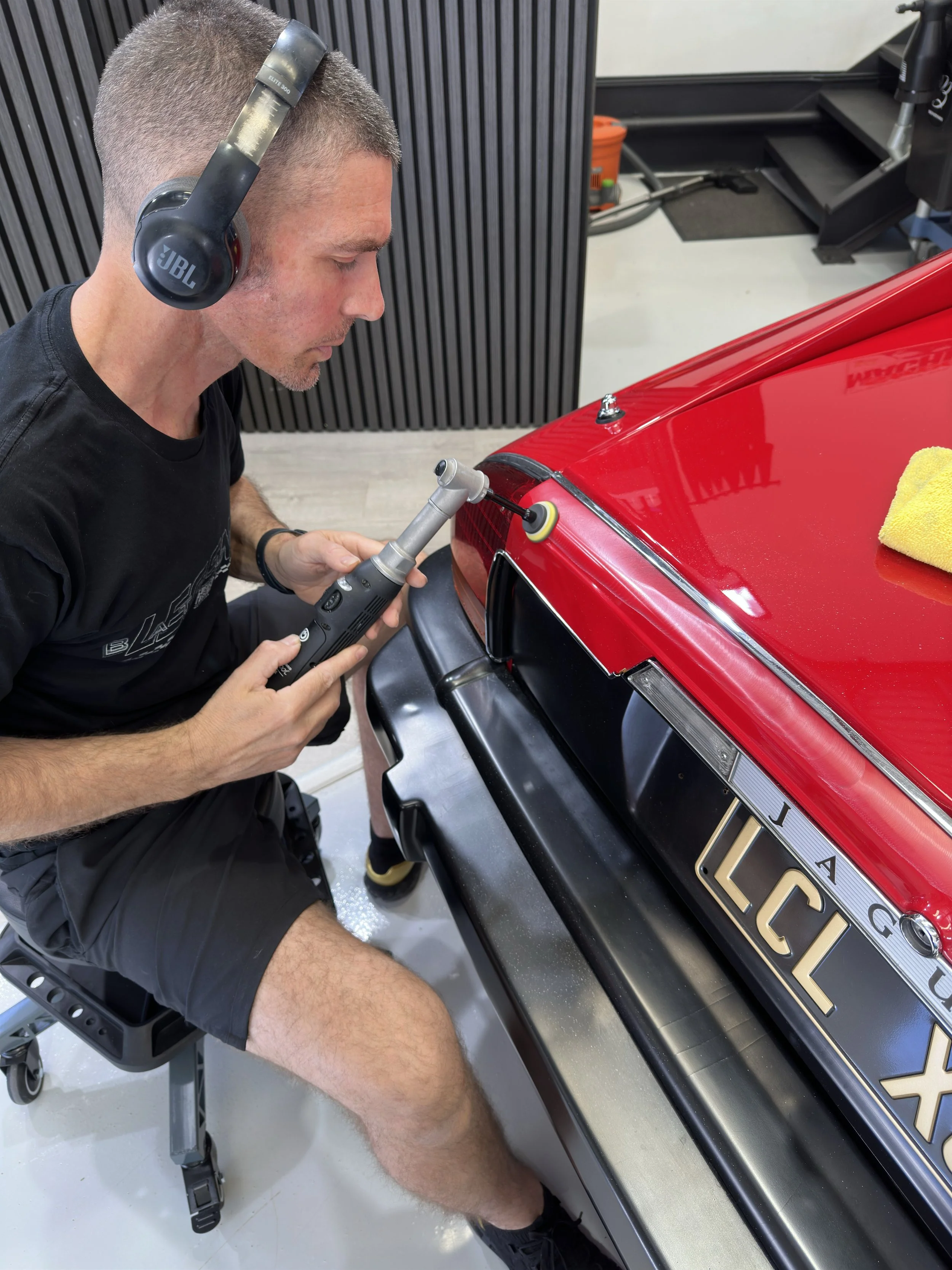 A man wearing headphones and black shorts is polishing the rear of a red sports car with a polishing tool. The car has a license plate that reads 'LUC LX' and a yellow towel is placed on the trunk. The setting appears to be a garage or workshop.