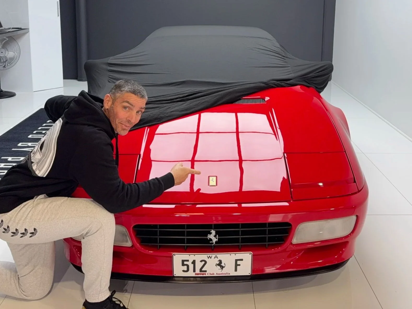 A man kneeling beside a red Ferrari sports car, pointing at a small emblem on the car’s hood, in a modern showroom with white tile flooring and a black cover over the car's rear.