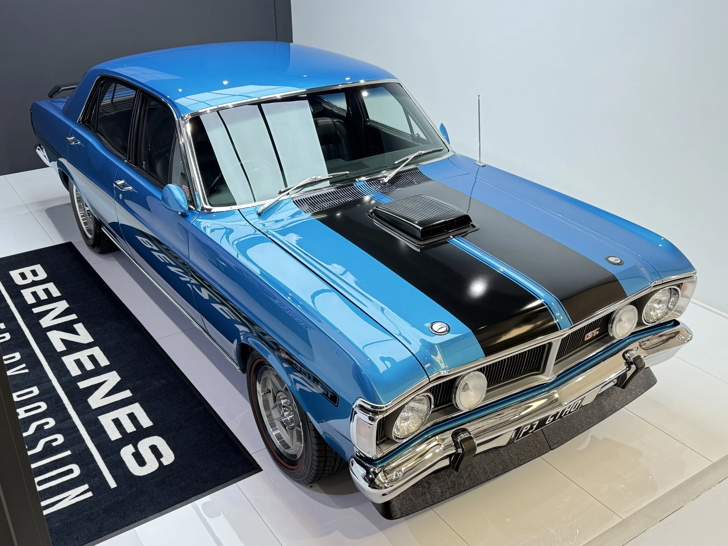 A classic blue and black race car on display. The car has a black hood, blue body with racing stripes, and a prominent hood scoop. It features round headlights, chrome bumpers, and racing decals on the side. The car is on a display mat that reads "Benzenes" and other text.