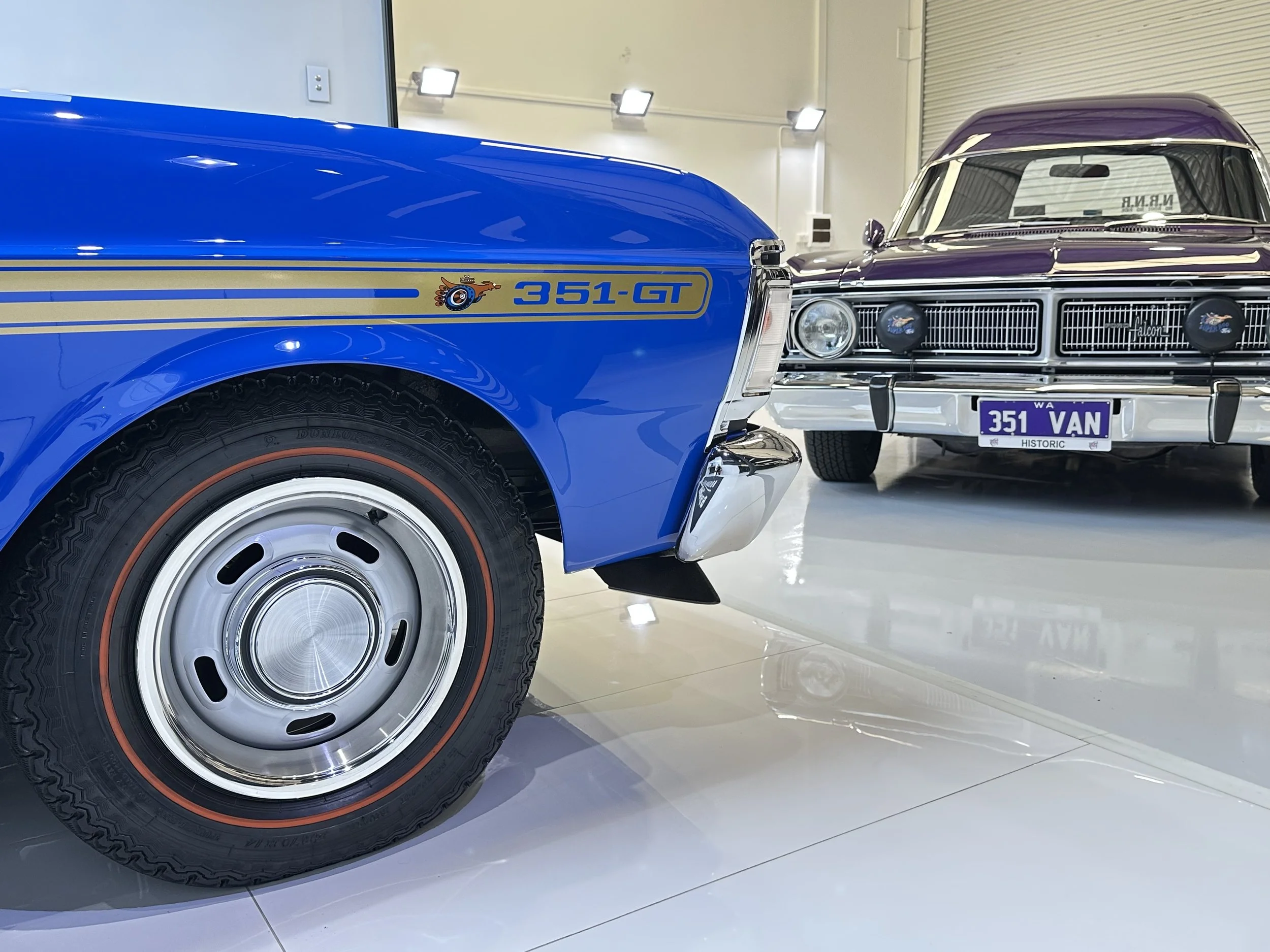 Close-up of a vintage blue Ford Falcon GT 351 car with a classic wheel, parked indoors with with another vintage car visible in the background.