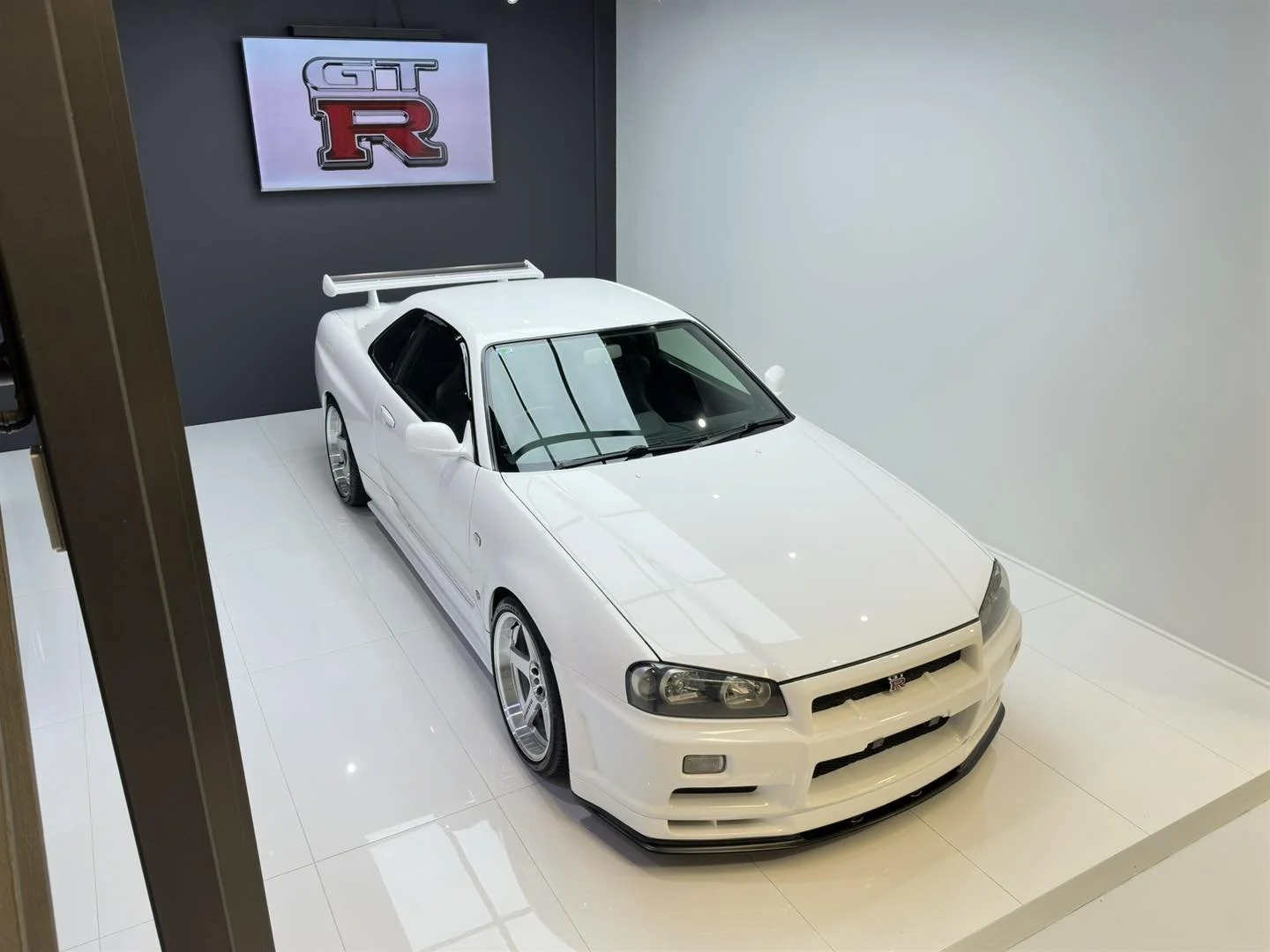 A white Nissan GT-R sports car inside a showroom, displayed in front of a dark gray wall with a GT-R logo on a display screen.