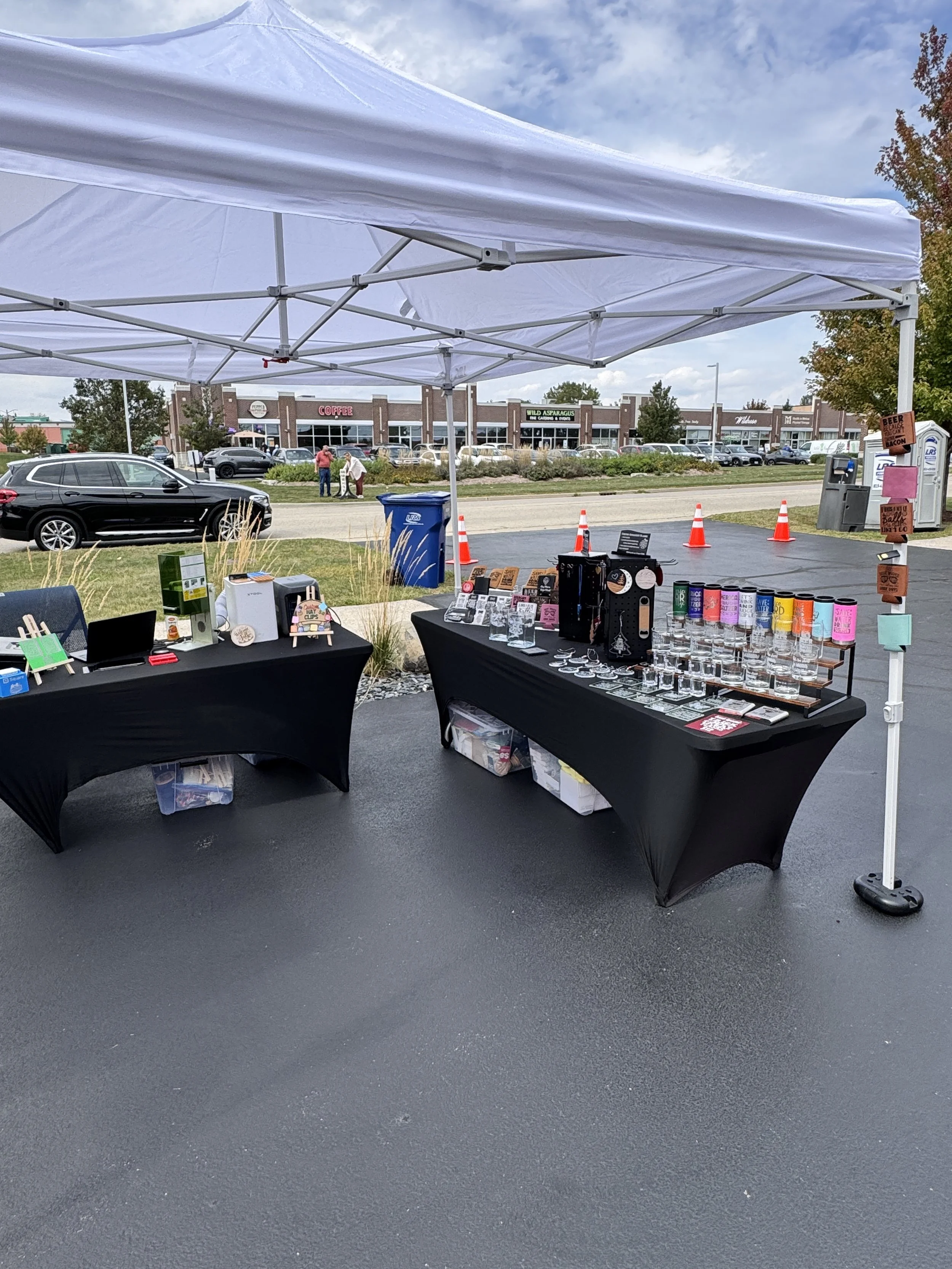 Outdoor booth setup with black tablecloths displaying various merchandise, under a white canopy in a parking lot, with stores and cars in the background.