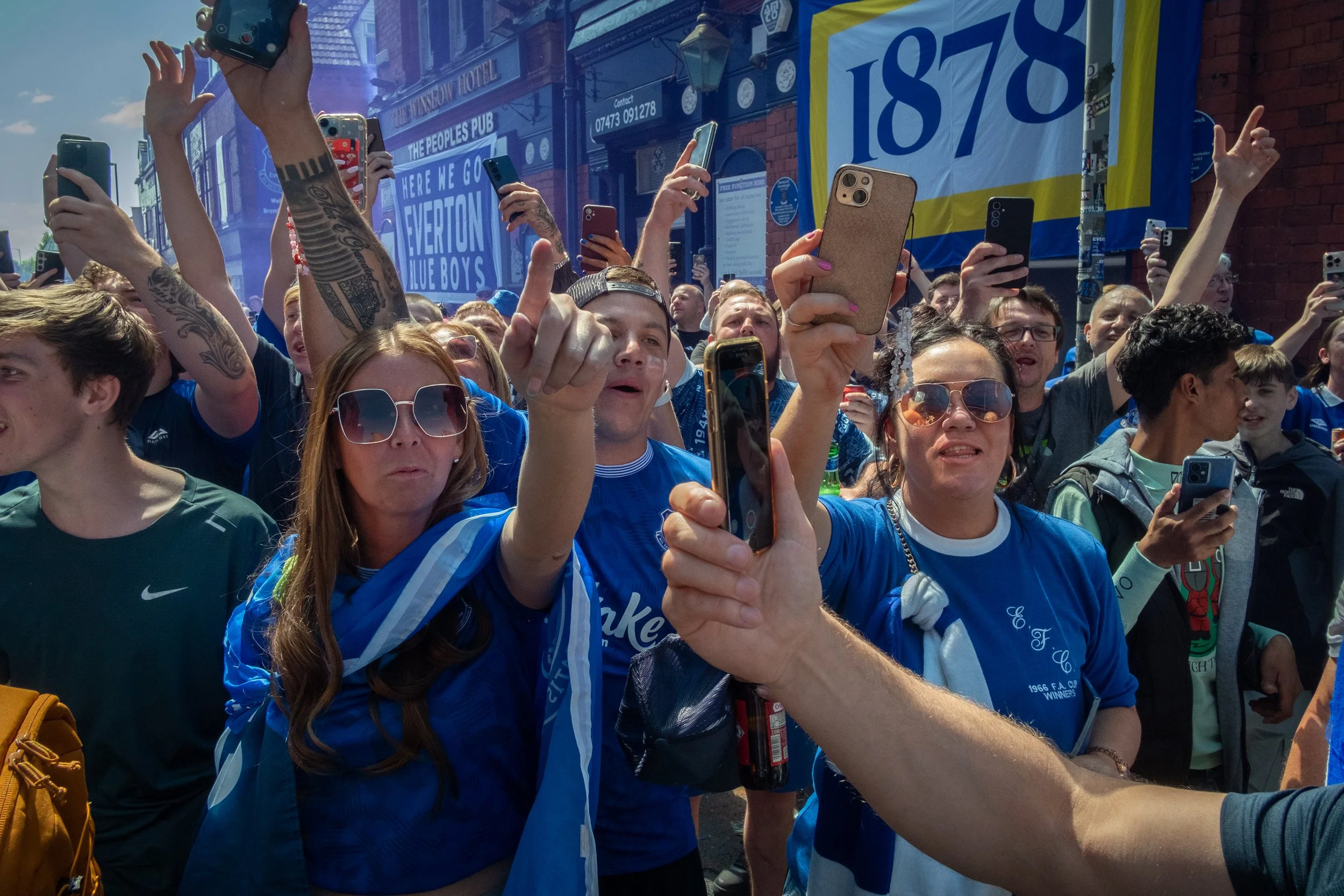 Crowd of people wearing blue shirts, taking photos and cheering at a sporting event outside.