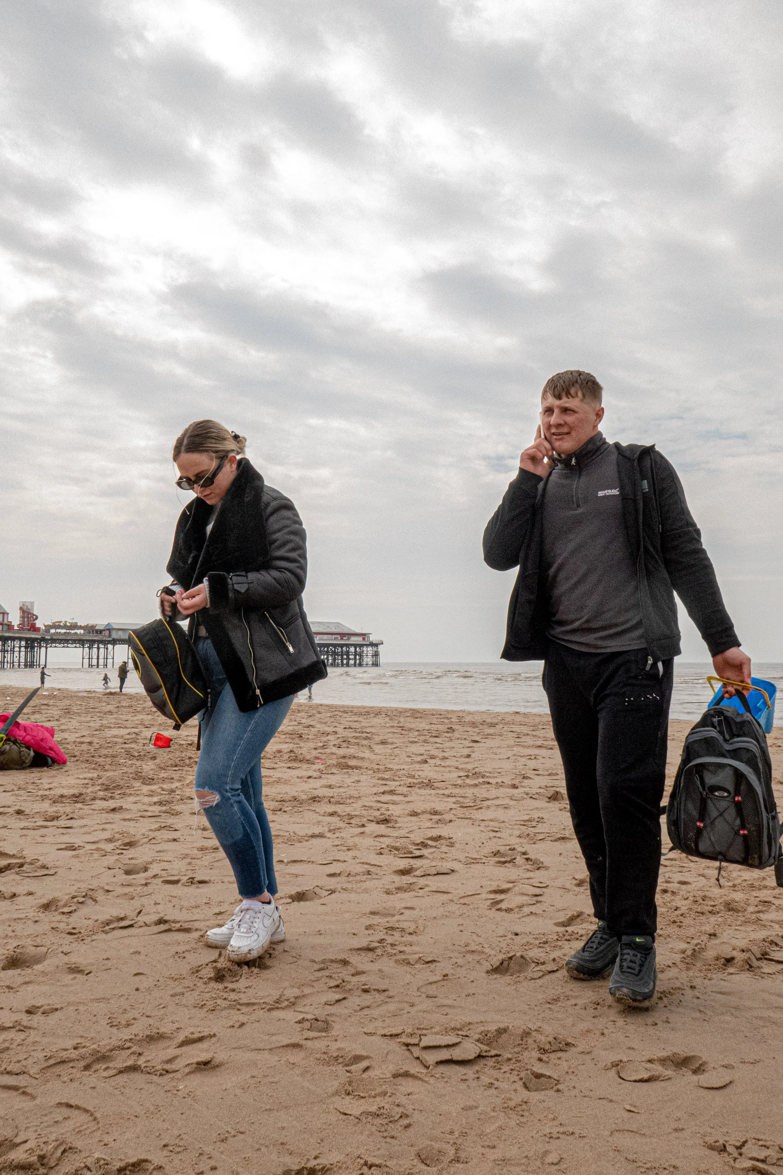 Two people walking on a sandy beach with a pier in the background. The woman on the left has long hair, wearing glasses, a black jacket, blue ripped jeans, and white sneakers, carrying a black bag. The man on the right has short hair, wearing a dark 
