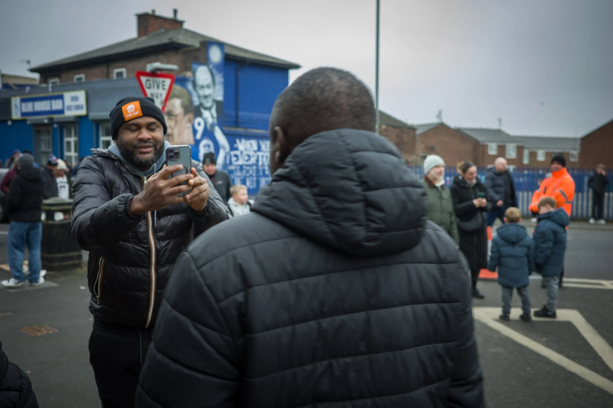 A man is taking a photo with his phone while standing next to another man in a black jacket. There are several people in the background, some wearing winter clothing, in an outdoor urban setting with buildings and graffiti murals visible.