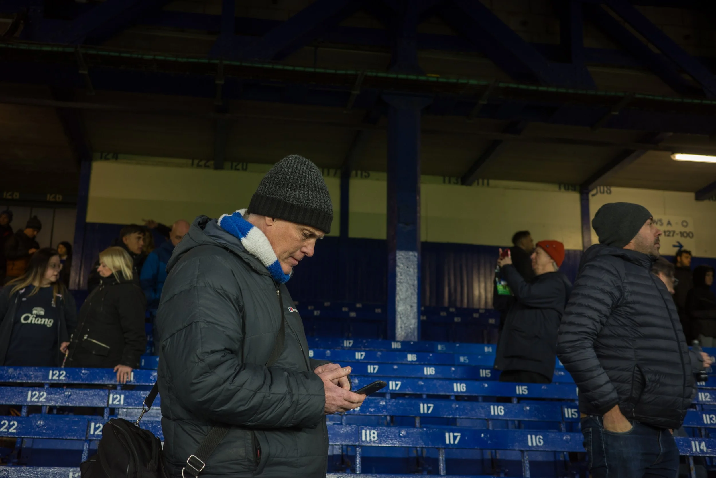 People standing in a stadium or arena, with blue seating, some using phones, one man in the foreground wearing a black knit cap and scarf, checking his phone.