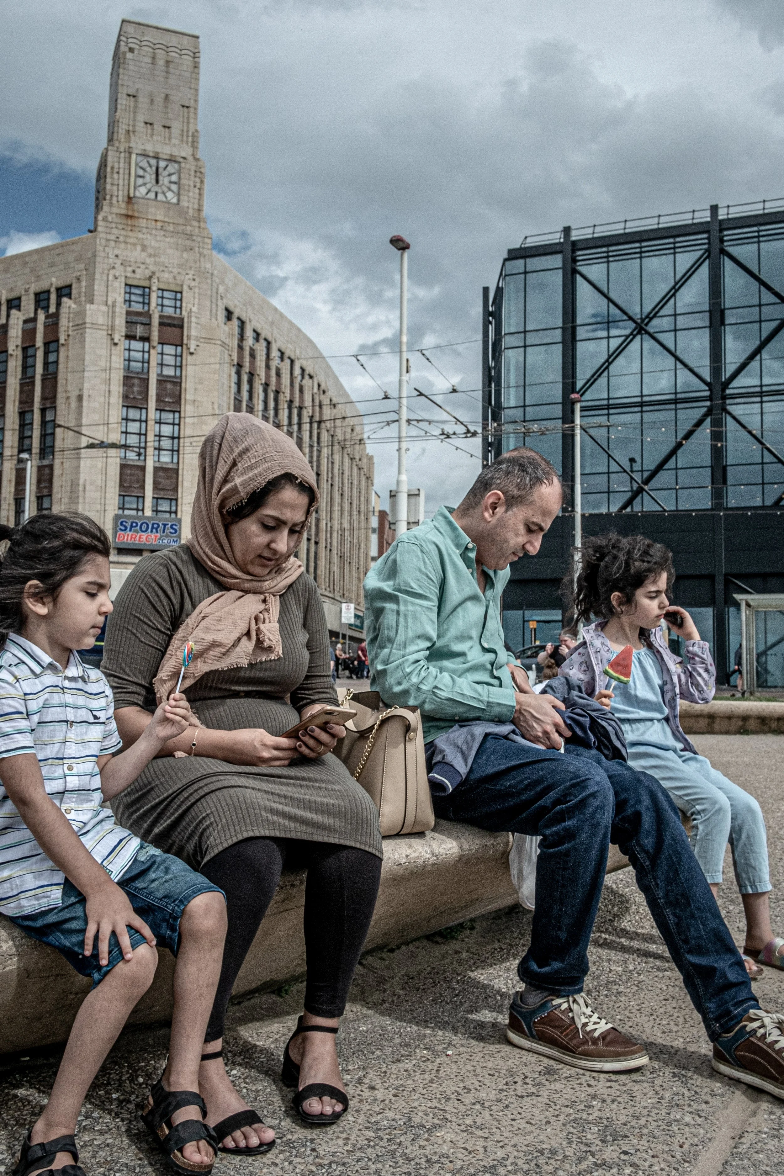 A group of people sitting on a bench outdoors in an urban area with tall buildings and cloudy sky in the background, some looking at their phones or holding small watermelon-shaped snacks.
