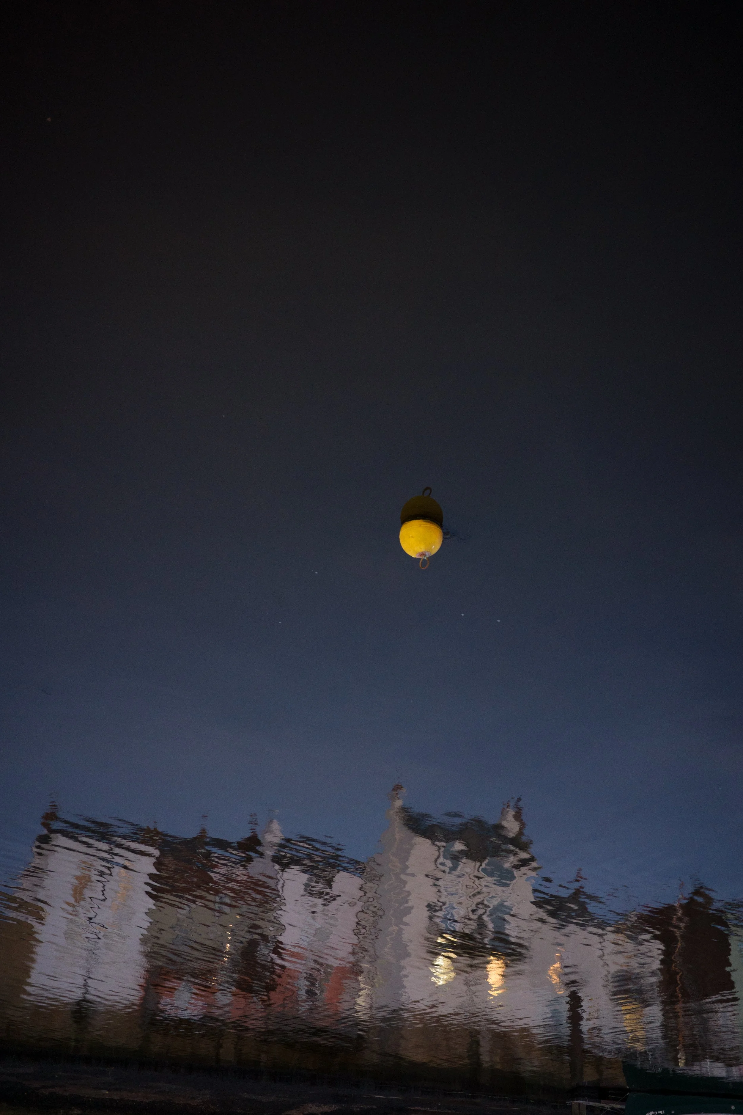 Reflection of a row of houses in water with a floating buoy or marker in the water and a dark sky above.