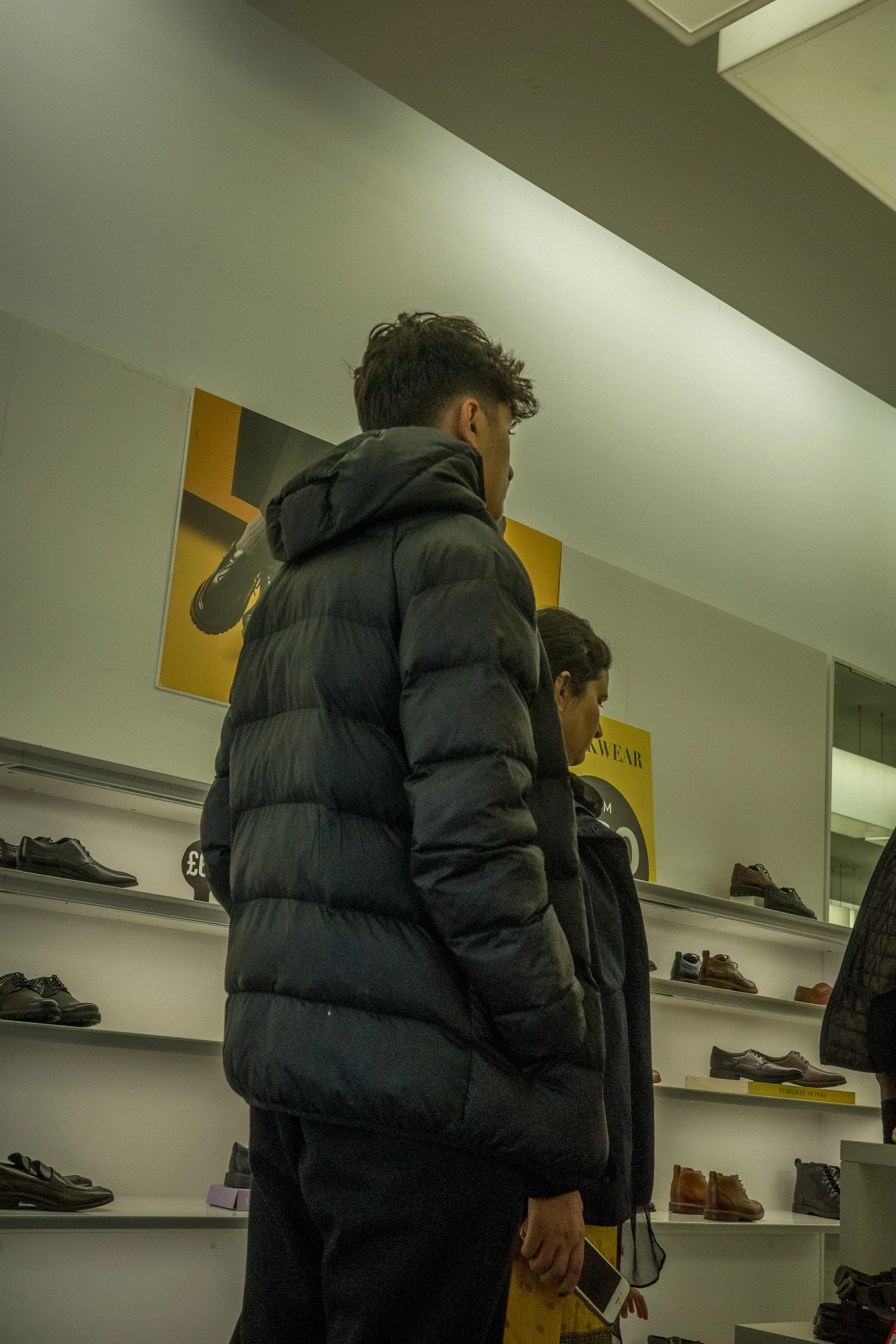 Two people shopping for shoes in a store, browsing a display of men's shoes on shelves.