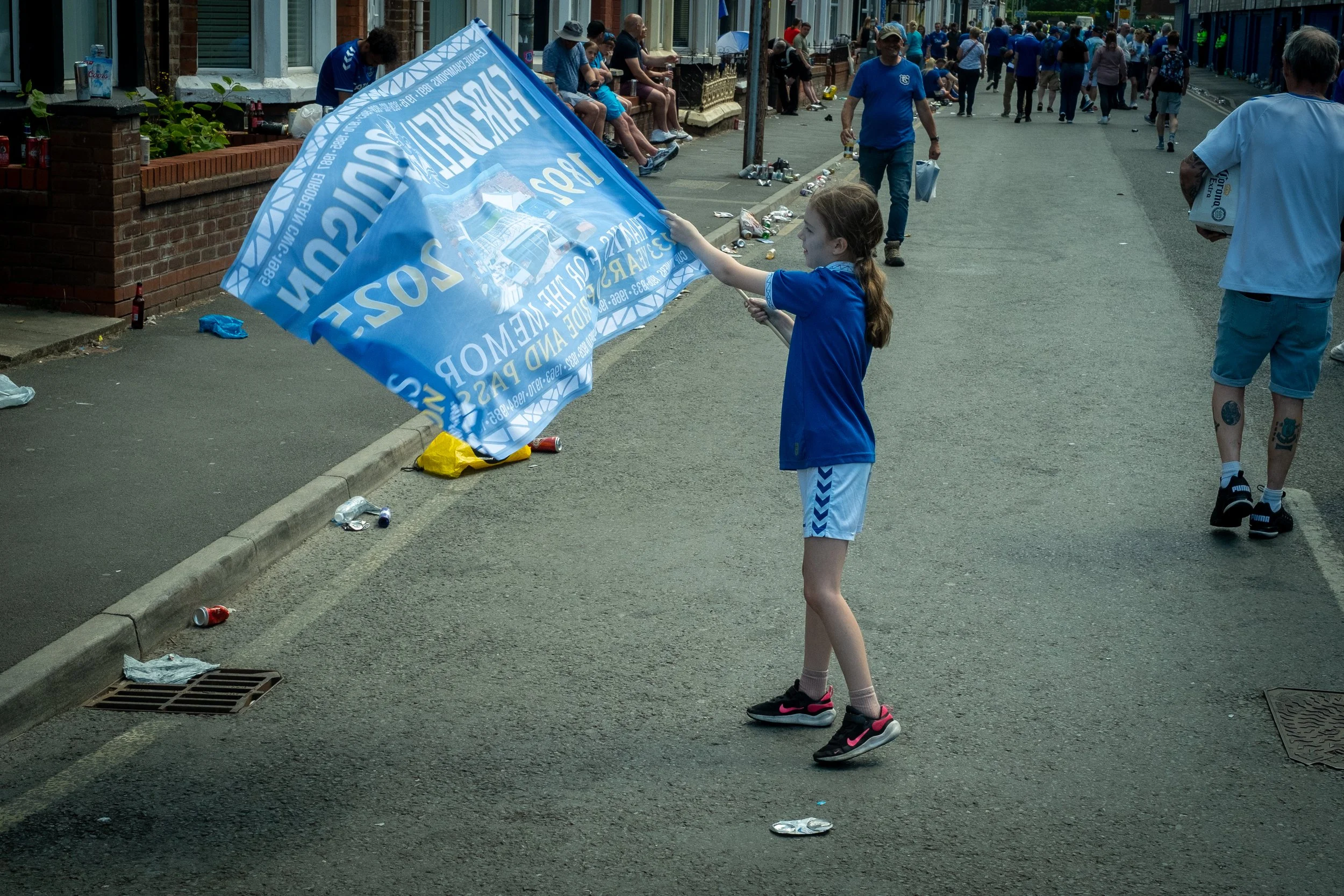 A young girl in a blue sports jersey and shorts is holding a large blue flag during a parade or street event, with many people sitting and walking in the background.