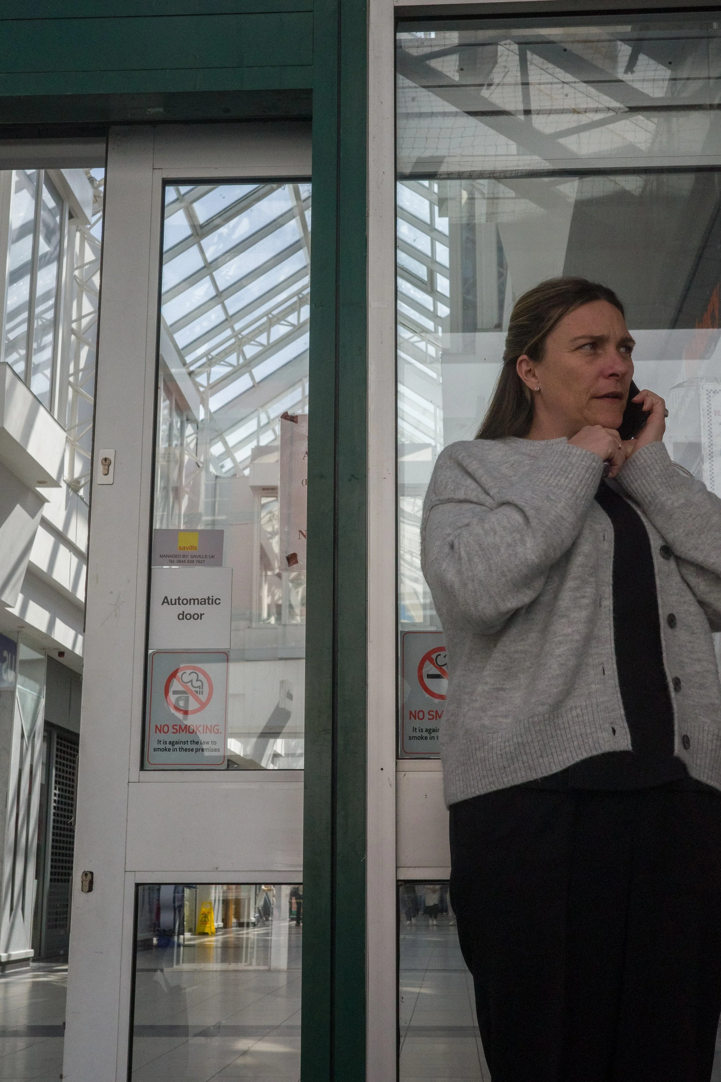 A woman with brown hair and wearing a gray blazer talking on a phone inside a modern building with glass walls and signs indicating no smoking and an automatic door.