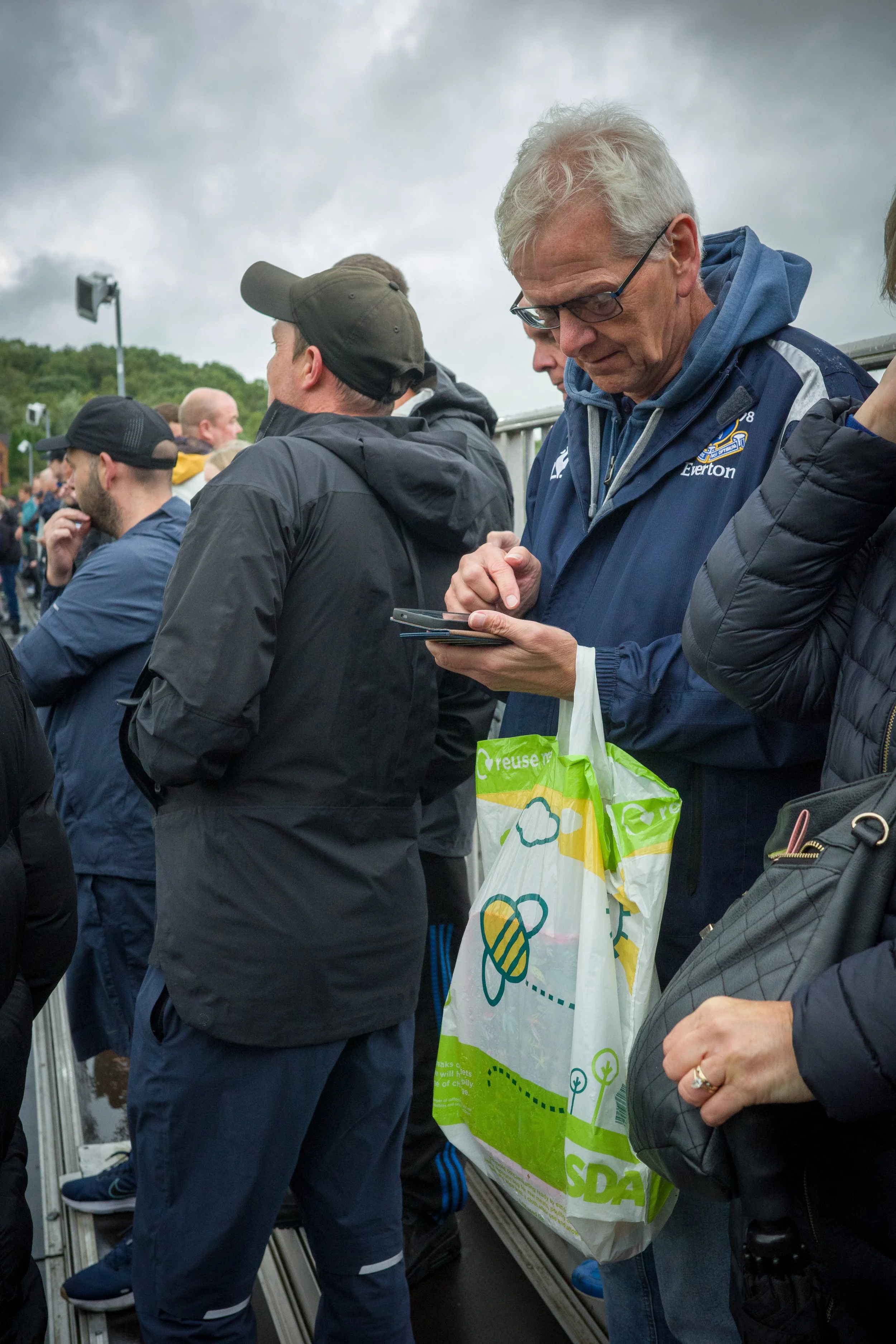 People standing in line outdoors on a cloudy day, some looking down at their phones, including an older man with glasses, gray hair, and a blue jacket, holding a reusable bag with green and yellow design.
