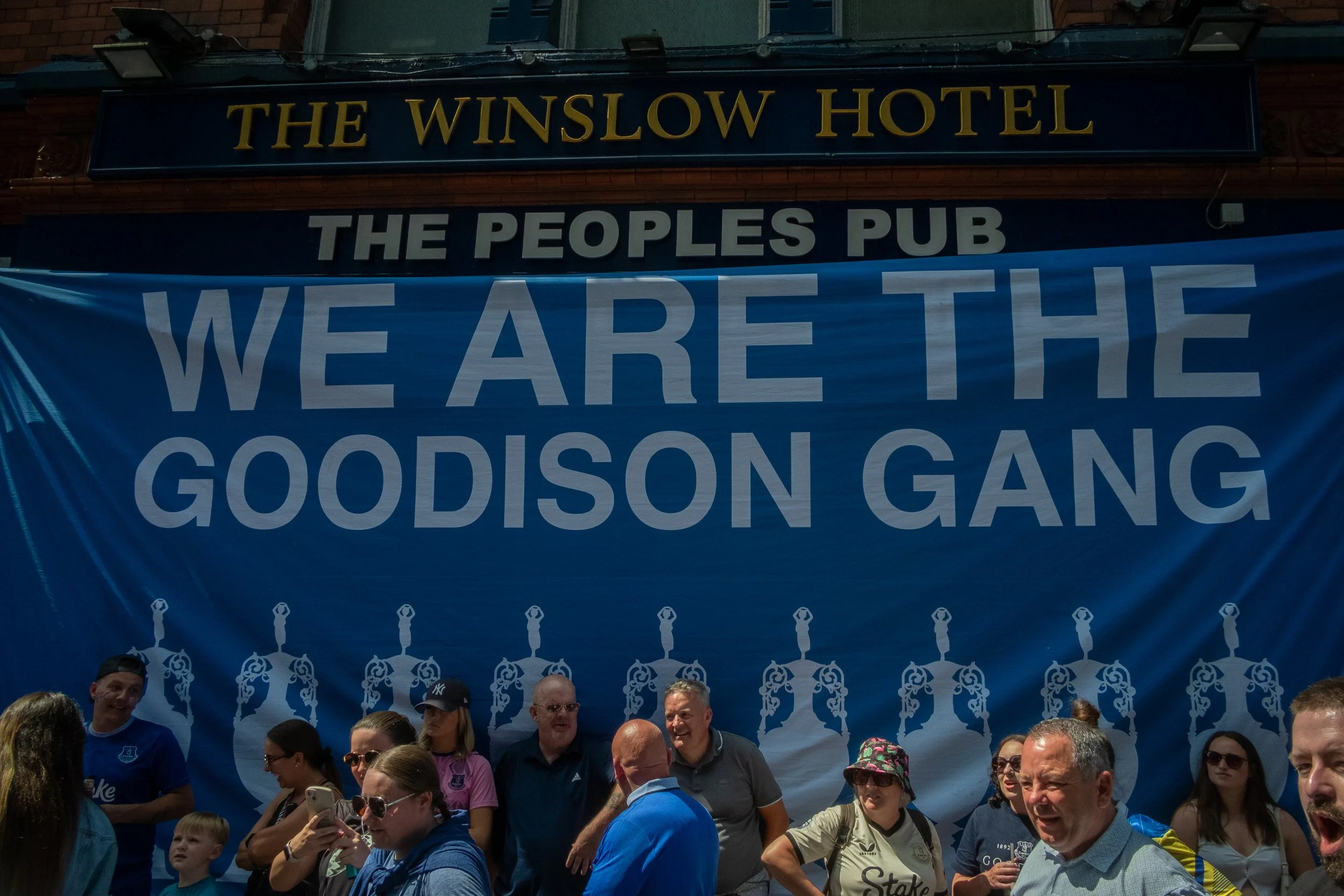 Group of people gathered in front of a large blue banner that reads 'We Are the Goodison Gang,' with the signs 'The Winslow Hotel' and 'The Peoples Pub' above the banner.