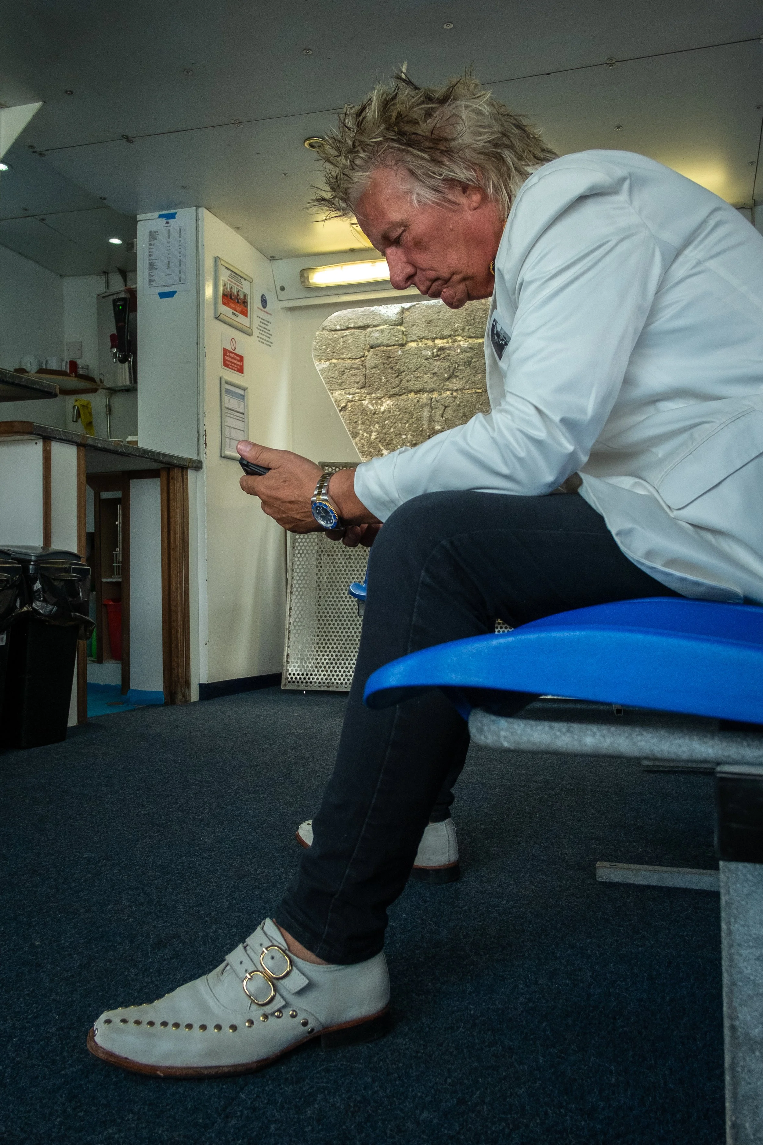 A person with wavy gray hair sitting on a blue chair, looking down at their phone, wearing a white jacket, black pants, and white shoes with gold accents and eyelets, inside a room with a gray carpet and white walls.