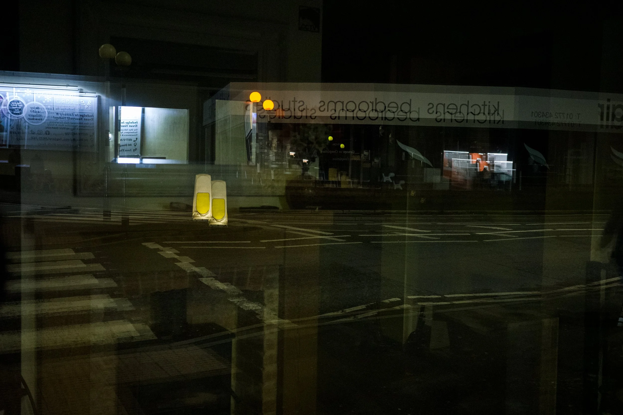 Nighttime view through a window of a parking lot with a few yellow traffic cones, some lit signage, and an illuminated interior space visible inside the building.