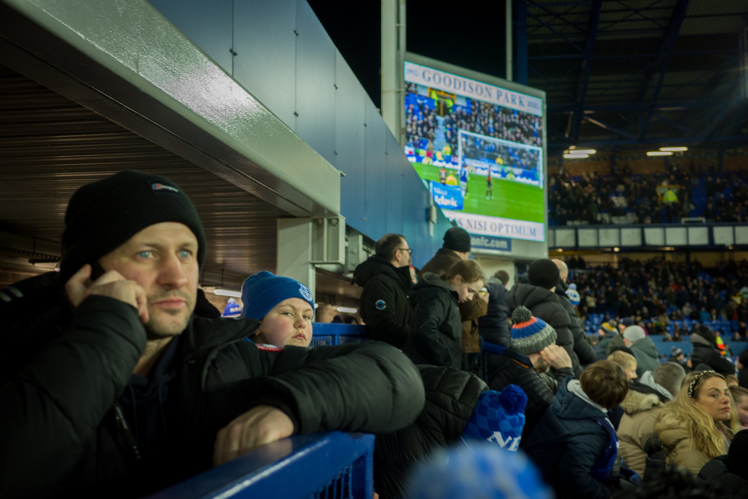 Crowd of spectators at a soccer game sitting in stadium seats, some wearing winter clothing and hats. A large digital scoreboard in the background displays the game.