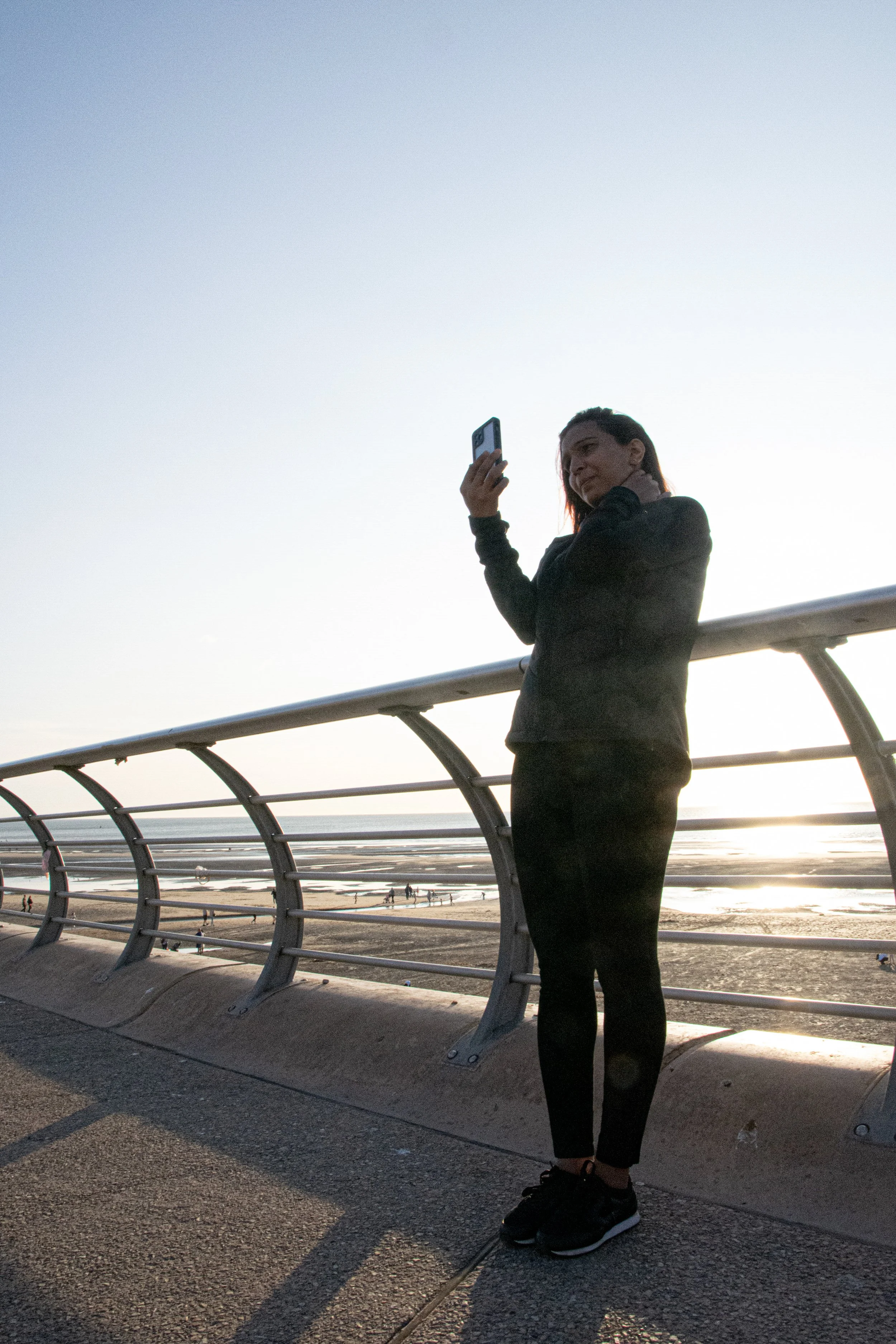 A woman in black athletic clothes and sneakers stands on an outdoor pathway, holding a phone, with a beach and ocean in the background during sunset, near a curved metal railing.