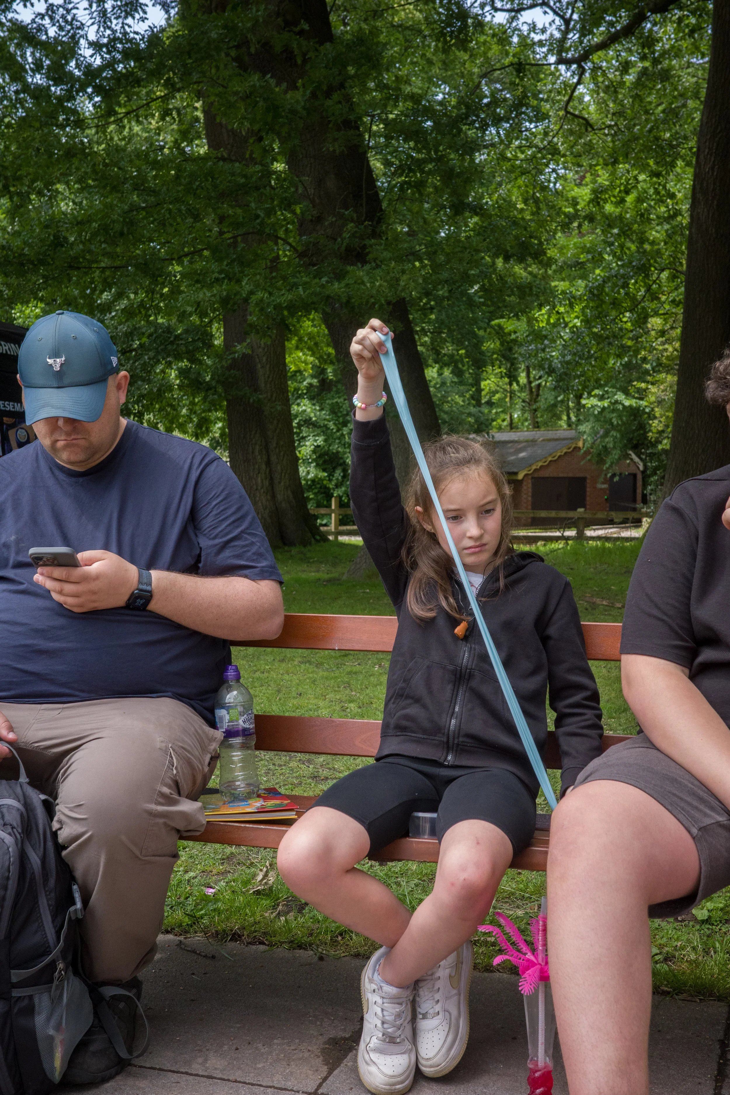 People sitting on a park bench surrounded by trees, with a young girl holding a long blue balloon. An adult man is looking at his phone, and a woman with curly hair is partially visible.