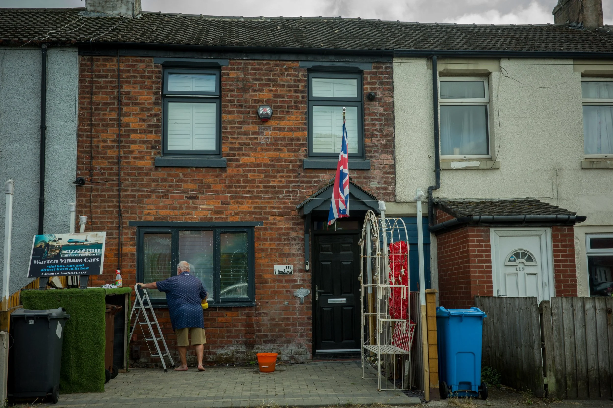 The image shows the front of a brick house with two windows and a small porch. A man with a ladder is cleaning or working on the house, with an older man standing nearby. There are various items in the yard, including trash bins, a sign advertising c