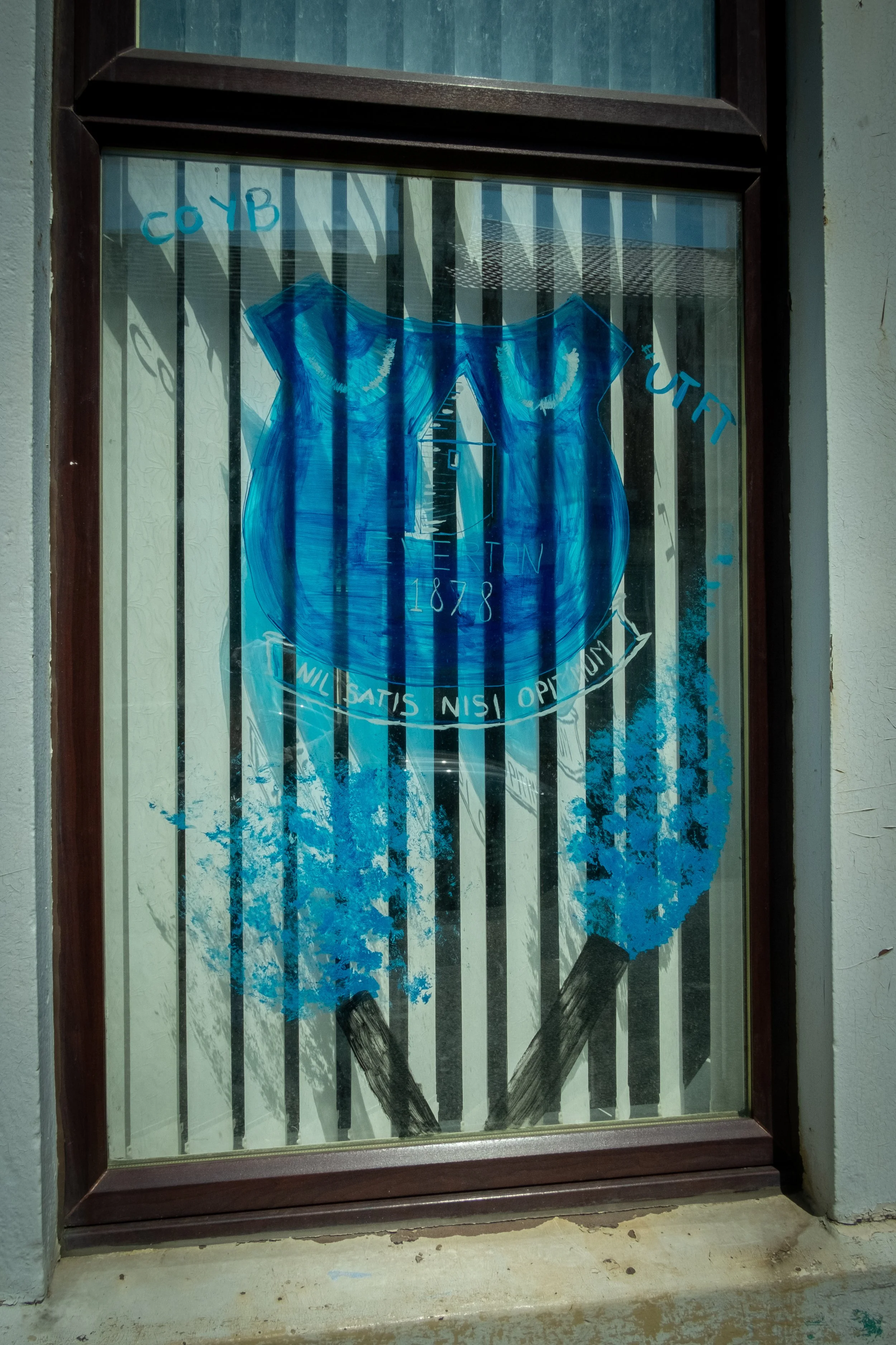 A painted shield on a window with blue and black paint, featuring the words "WILL SATIS NISI OPTIMUM" and the date 1878, with a background of black and white vertical stripes.