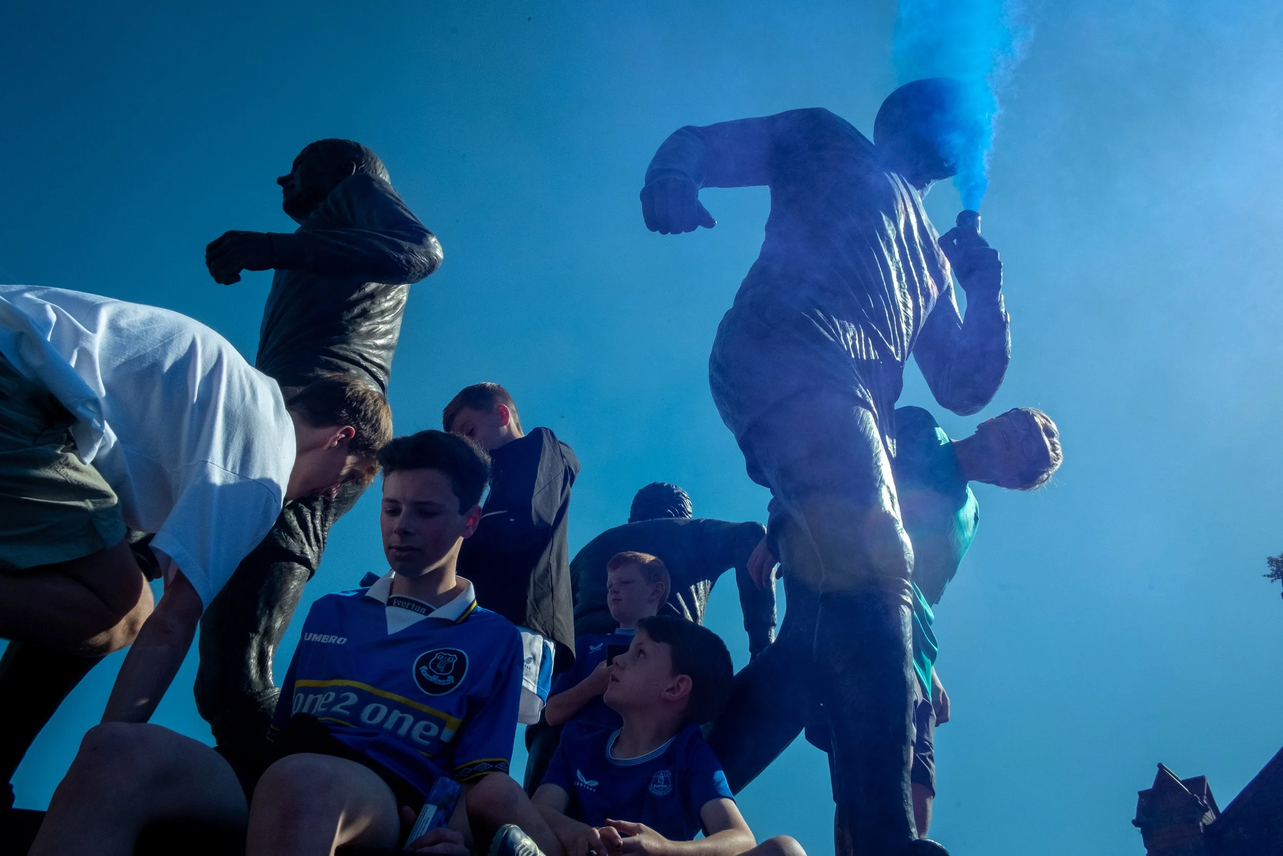 People, including children and teenagers, gathering around statues of football players, some sitting and others standing, with a person holding a smoking flare, against a bright blue sky.