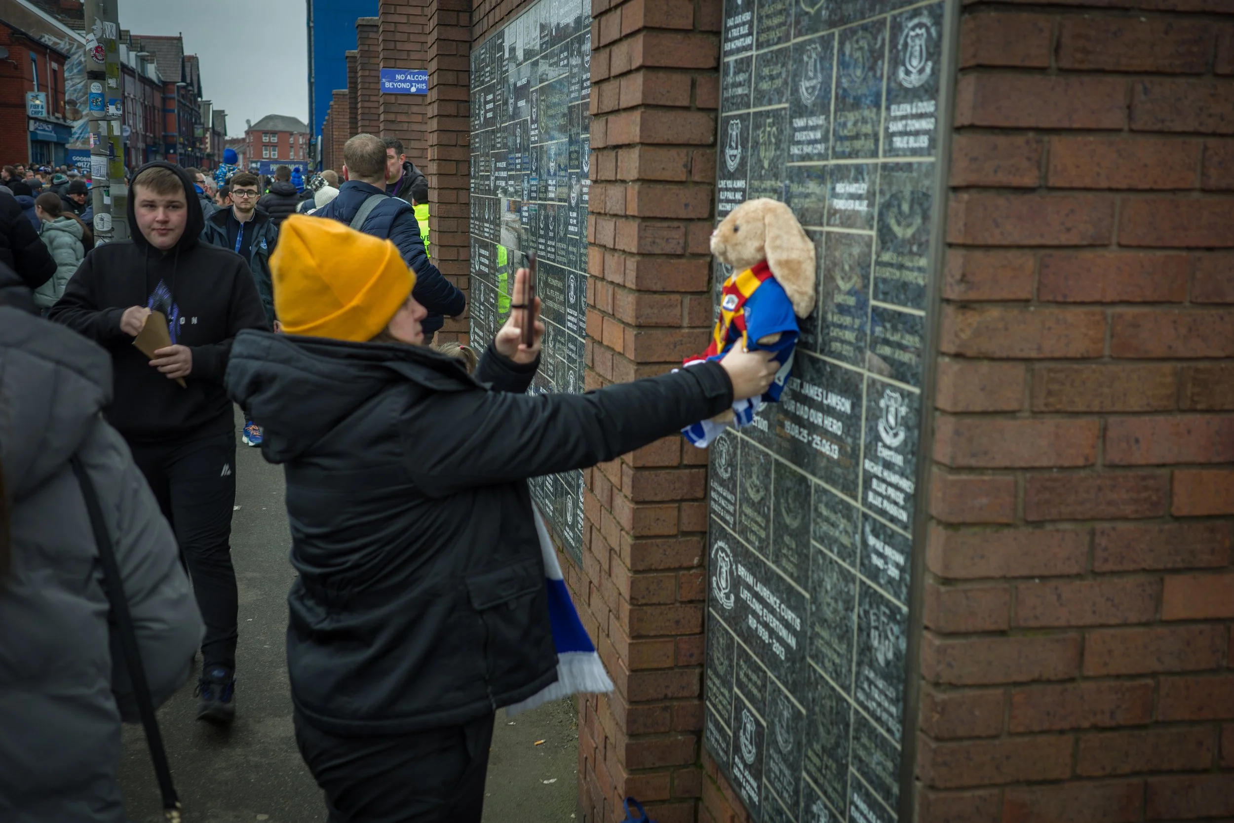 A crowd of people gathered around a memorial wall with plaques, with one person placing or adjusting a stuffed animal dressed in a colorful sports jersey on the wall. The memorial wall has numerous plaques, likely commemorating individuals or events,