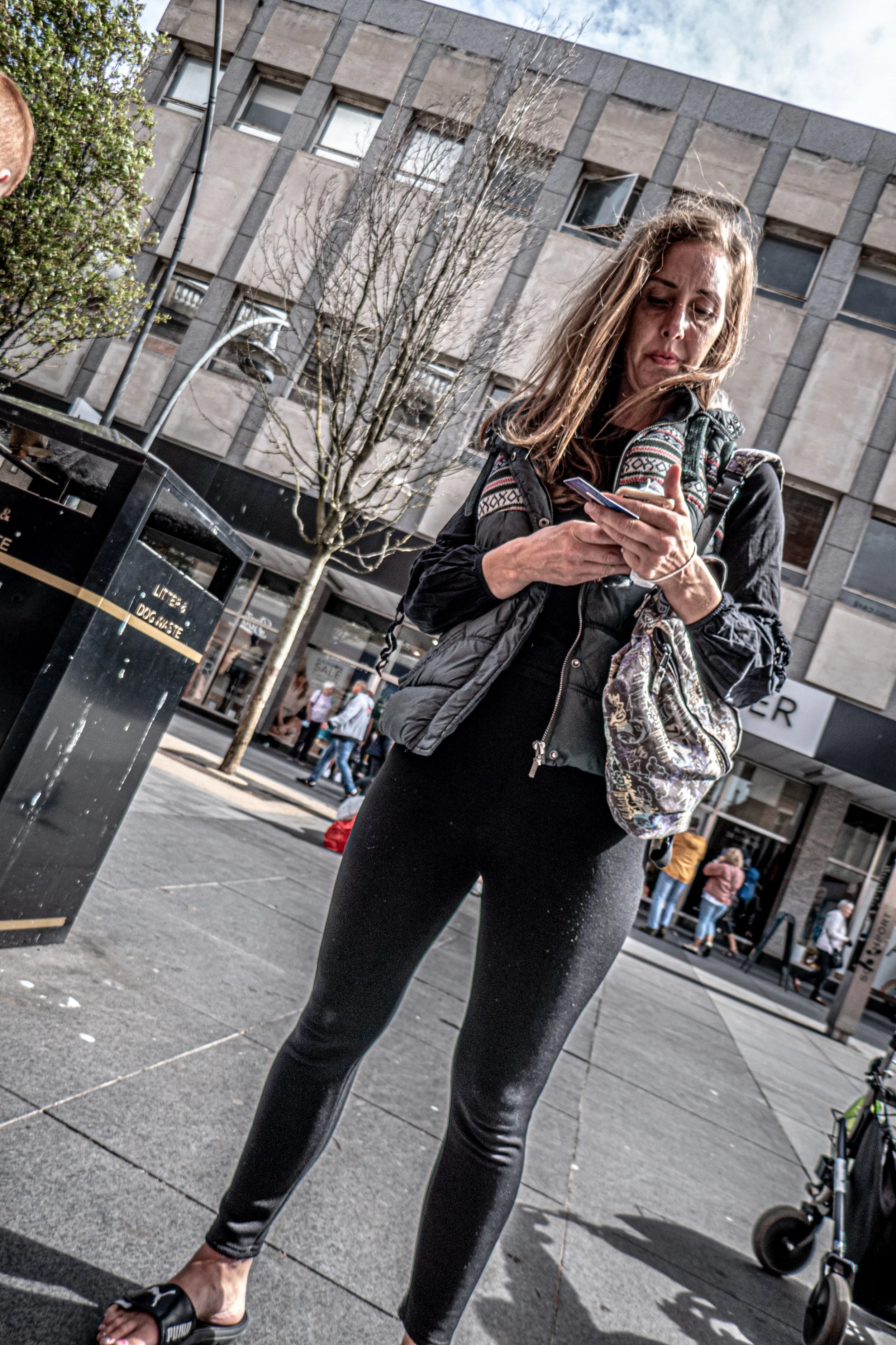 A woman with long, light brown hair is standing outdoors on a city street. She's wearing a black jacket, black leggings, and black sandals, and is looking at her phone. In the background, there are buildings, a leafless tree, several pedestrians, and