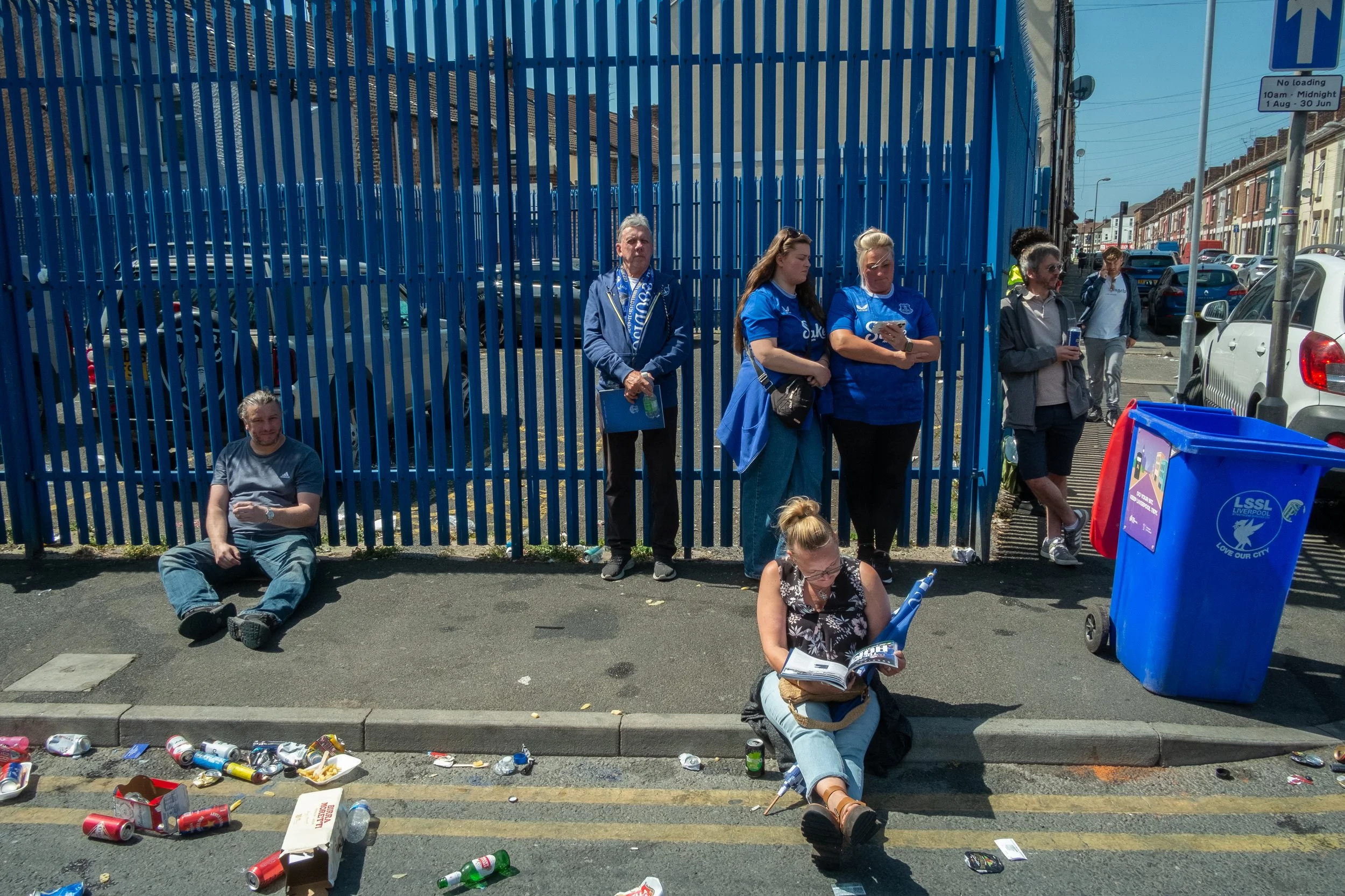 People standing and sitting on the sidewalk outside a blue fence, surrounded by litter including cans and food containers.
