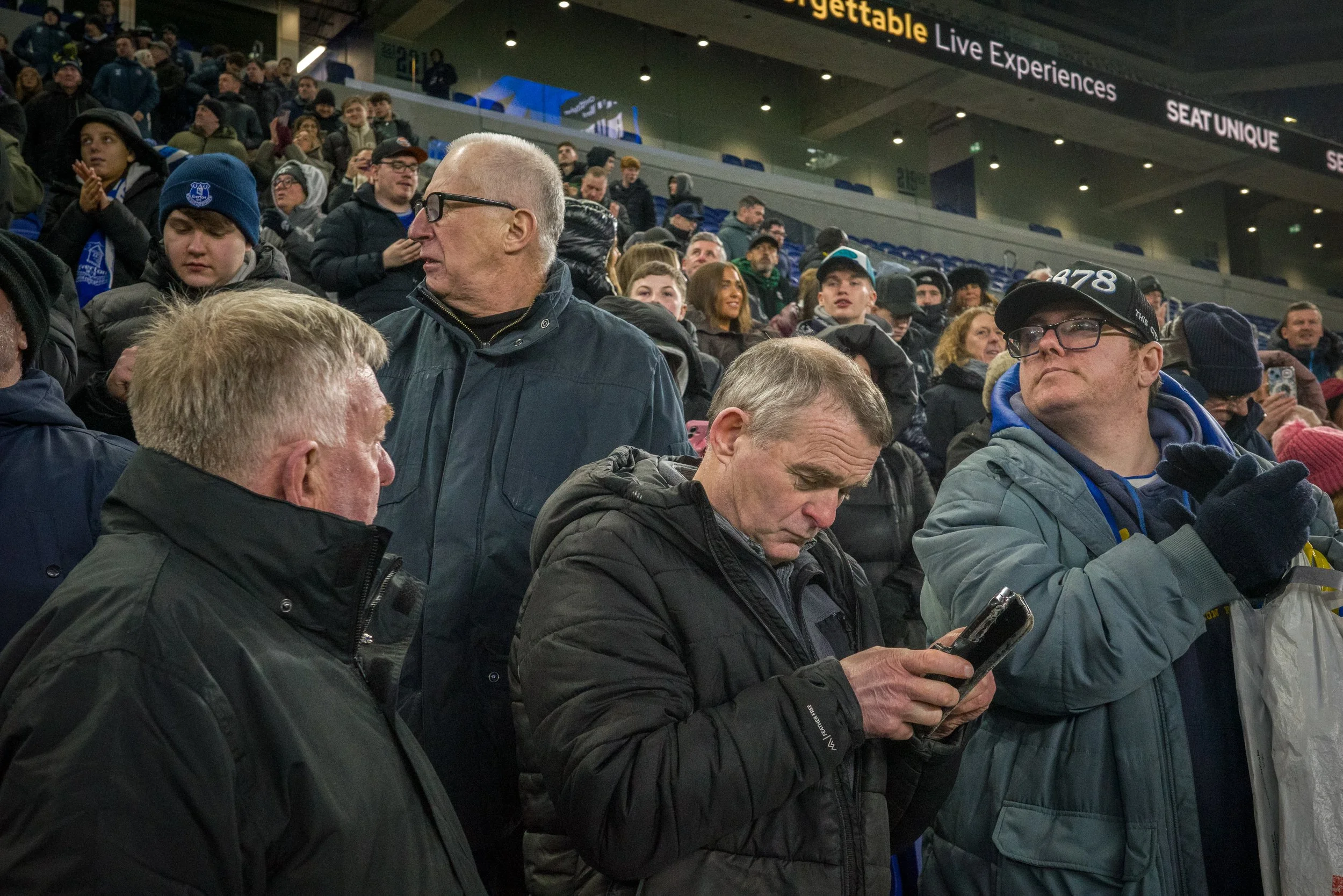 Crowd of people sitting inside a stadium, some looking at their phones, others talking, wearing winter clothing, with advertisements and signs visible in the background.