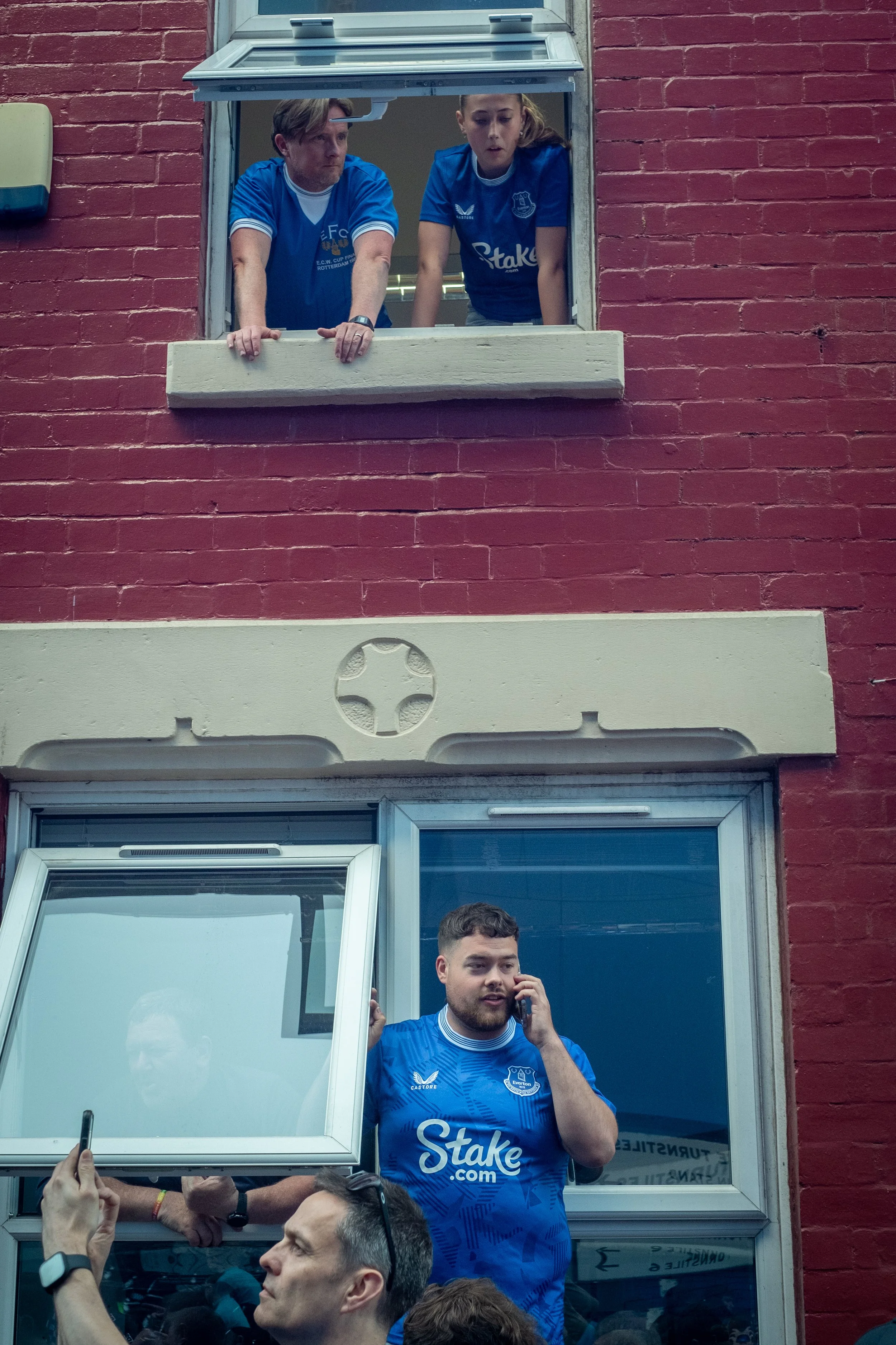 Two people looking out of a window on the second floor of a red brick building, and a man in a blue sports jersey talking on a mobile phone outside the building.