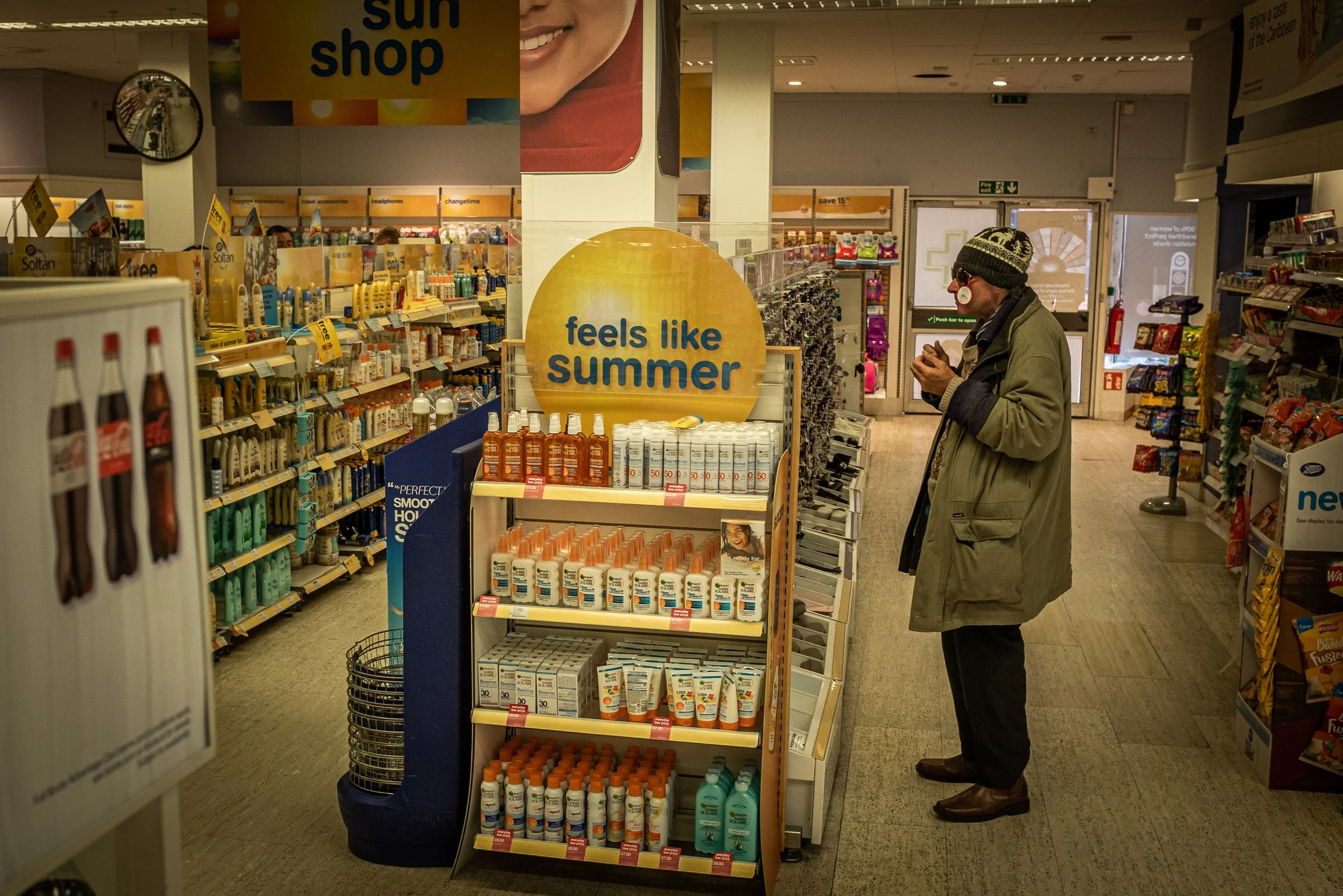 A man in a green coat and knit hat standing in a pharmacy aisle looking at products. The aisle has a yellow sign that says 'feels like summer' and is stocked with sunscreen and sunblock products. Shelves on the right display snacks and other items.