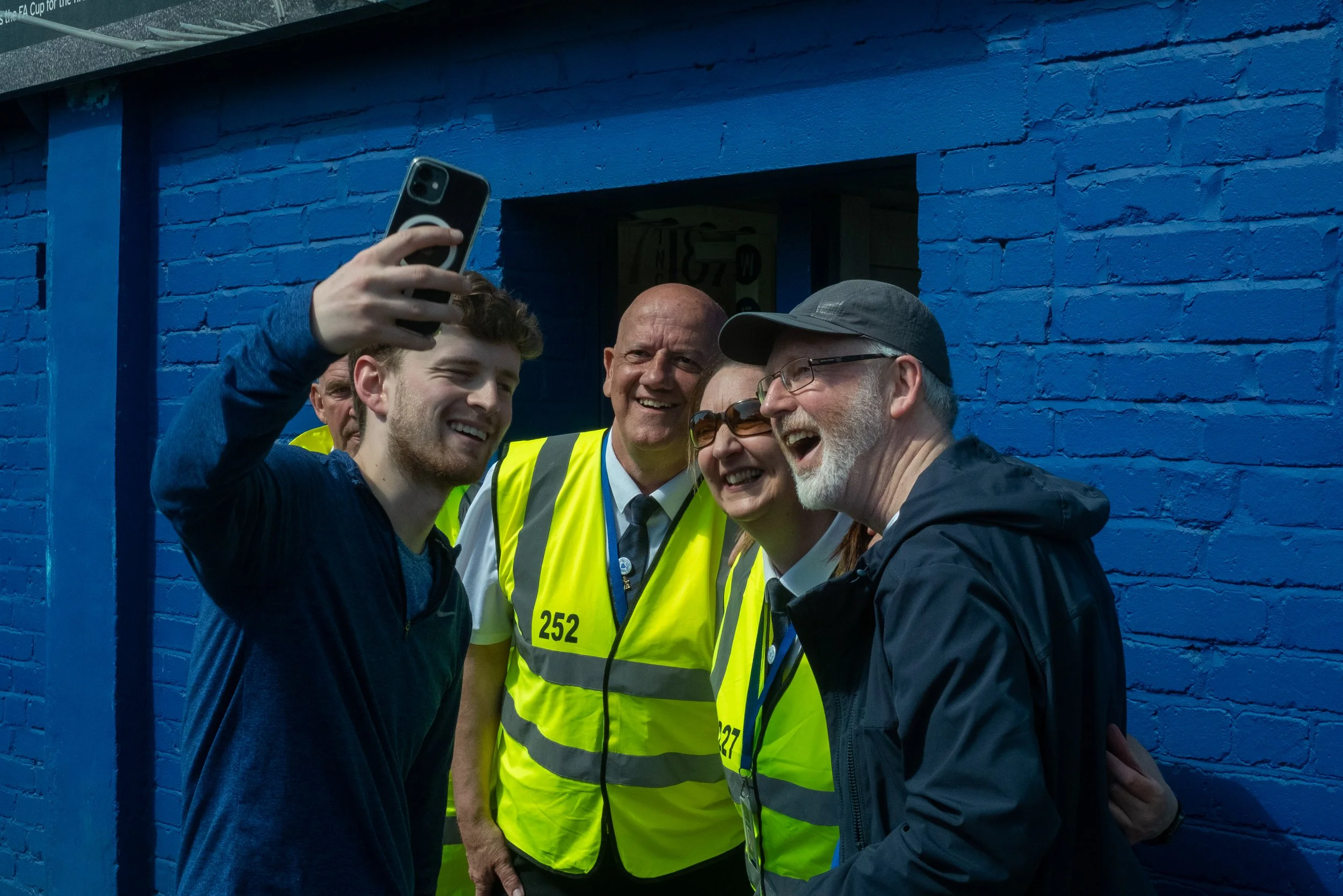 Four smiling people taking a selfie in front of a blue painted brick wall. Two are wearing yellow safety vests with numbers, and the others are dressed casually, one with a cap and glasses.