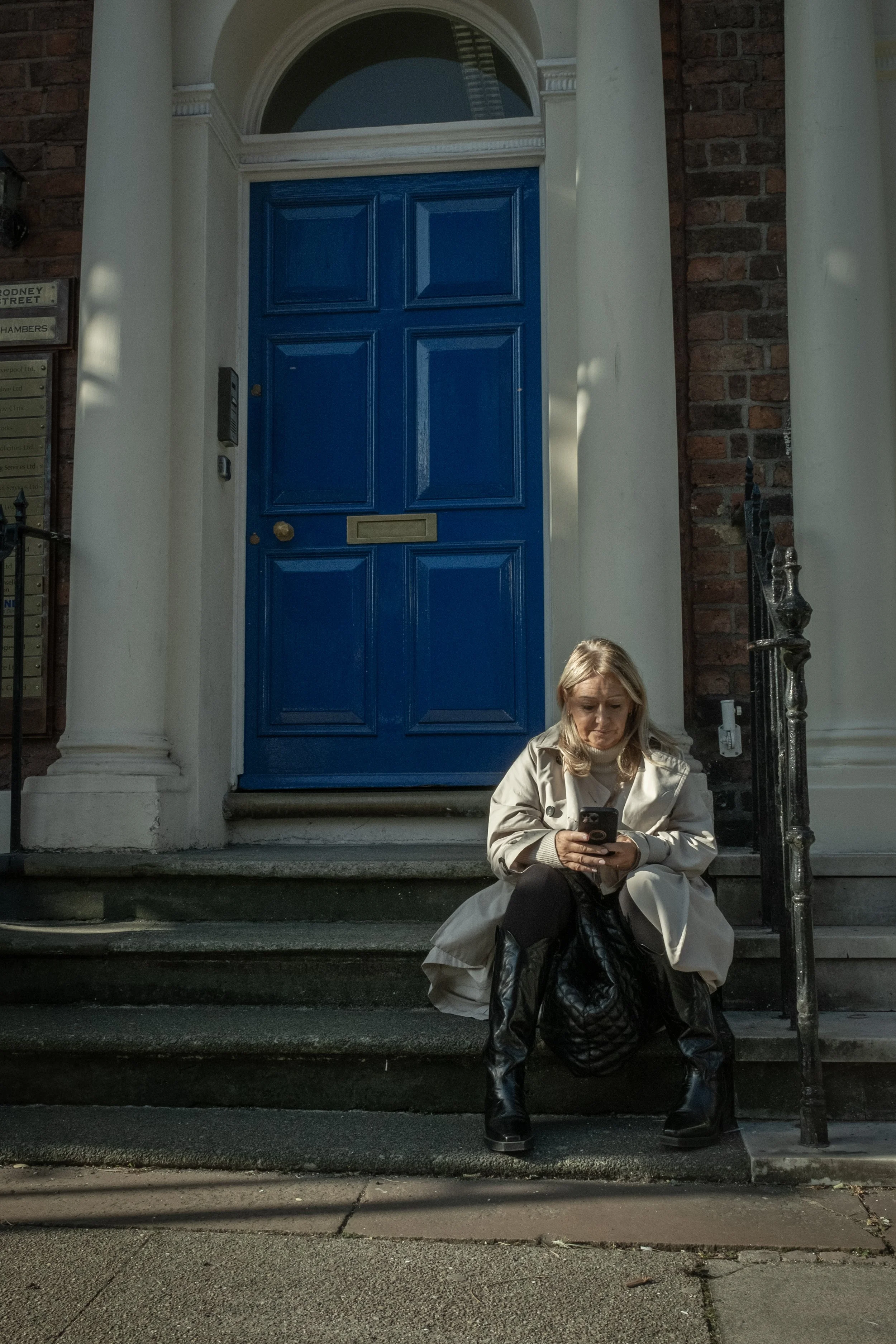 A woman with blonde hair is sitting on the steps of a building with a bright blue door, using her phone. She is dressed in a beige coat and black boots, with a black handbag placed in front of her. The building has brick and white walls, with decorat