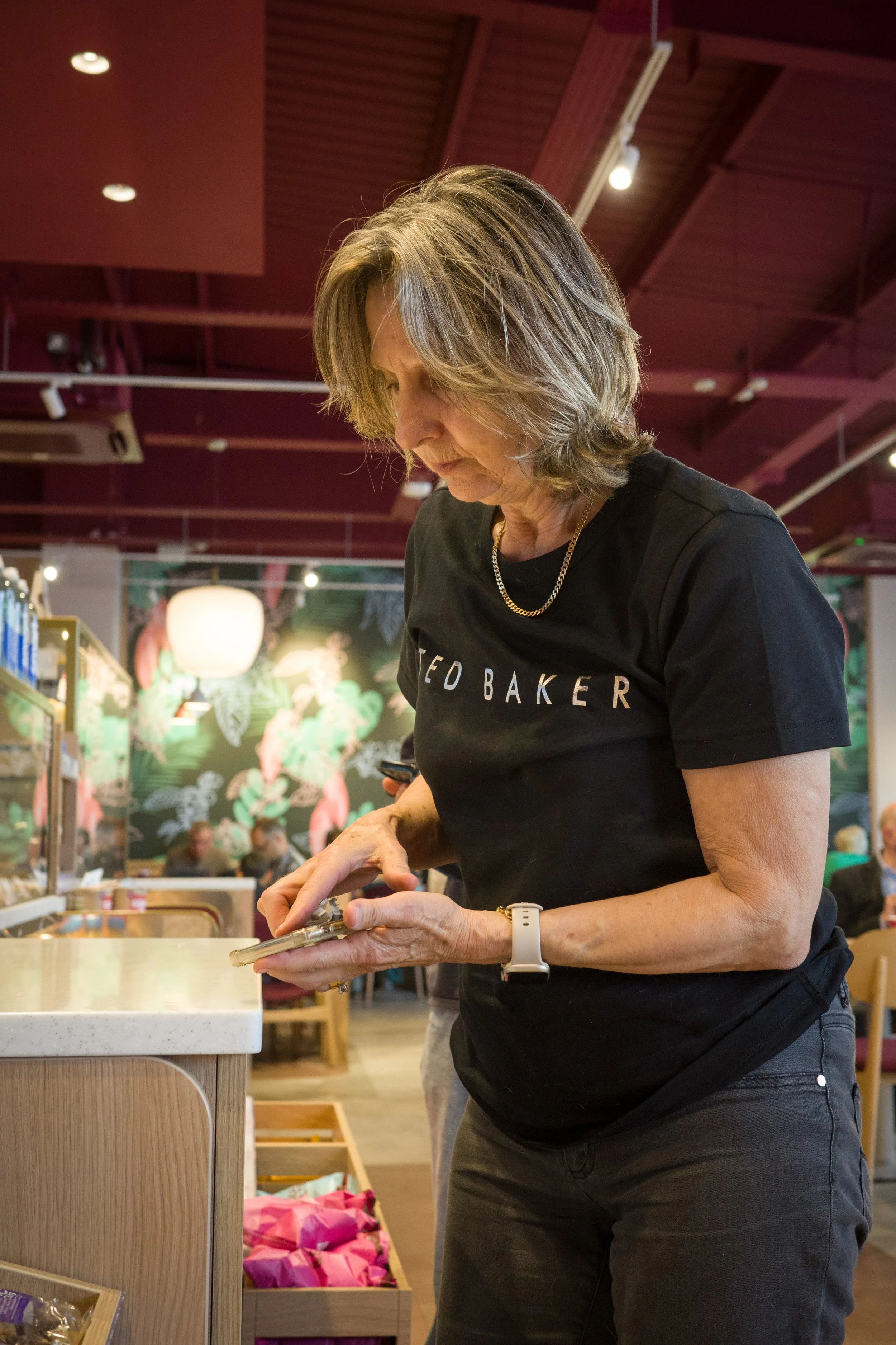 A woman with shoulder-length hair wearing a black t-shirt with white text, looking down at her phone in a store with a red ceiling and colorful wall art in the background.