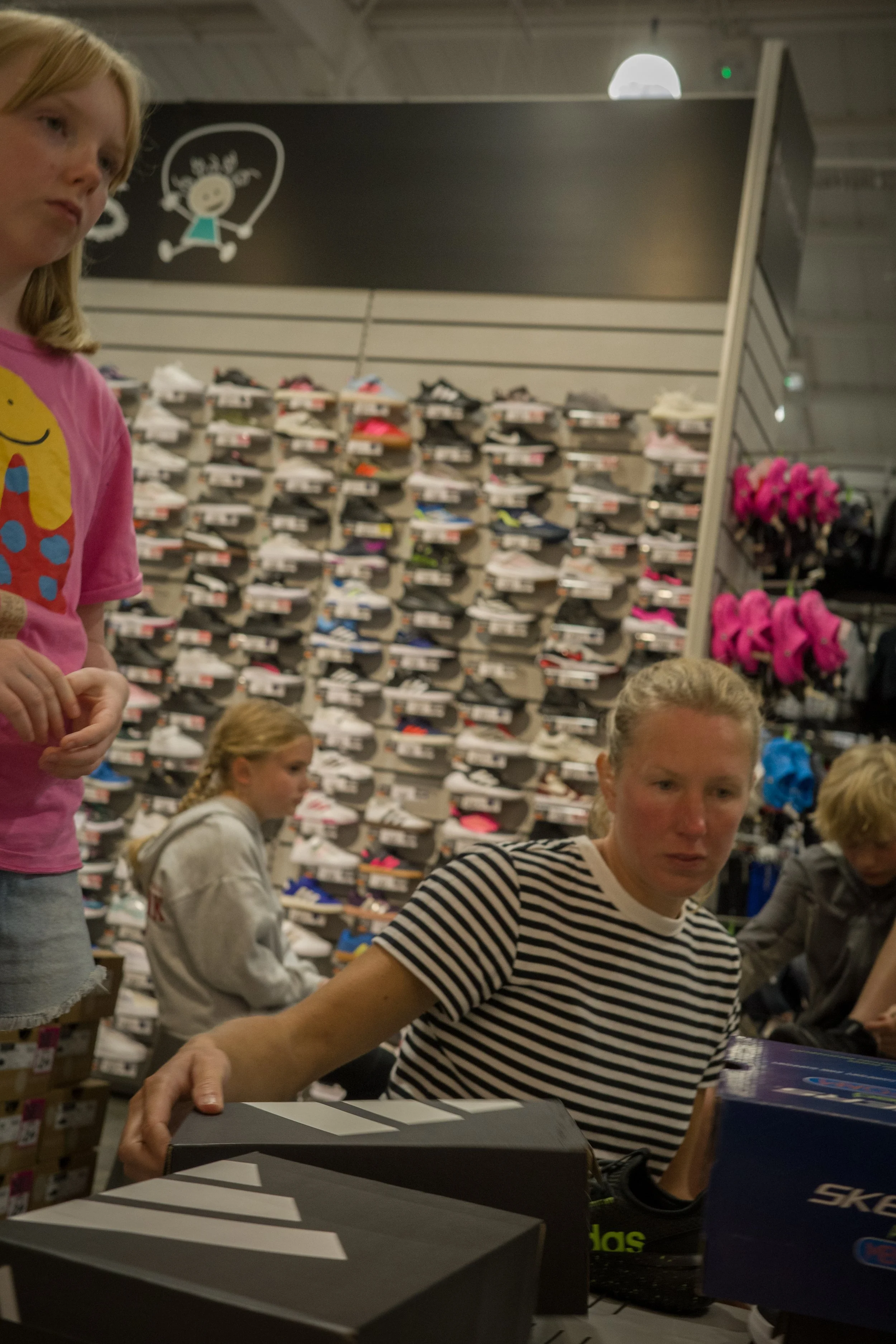 A woman wearing a striped shirt shopping for shoes in a retail store, with multiple shelves of sneakers in the background.